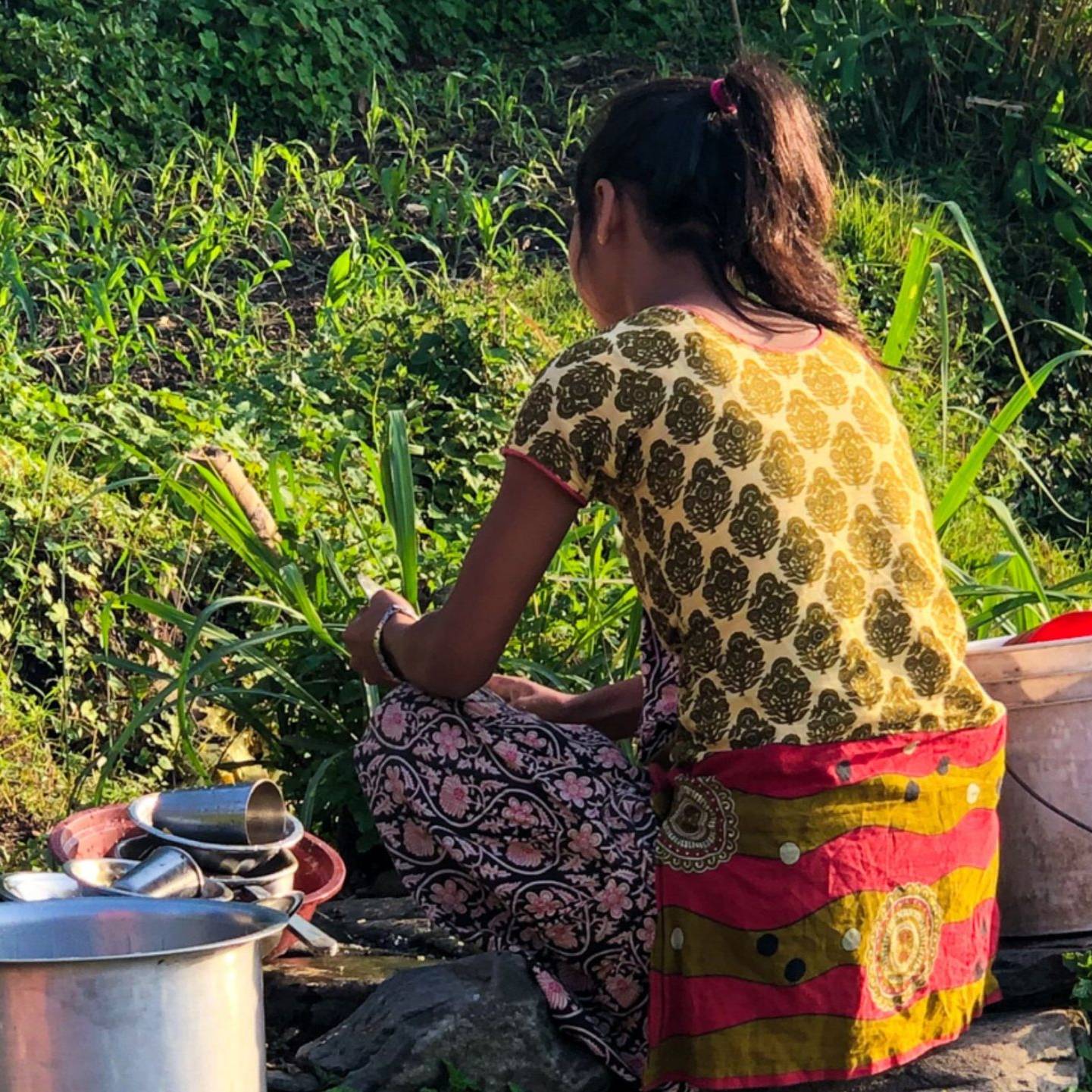 A young woman sits outdoors, surrounded by lush greenery, washing dishes with buckets and utensils by her side, illuminated by the warm sunlight.