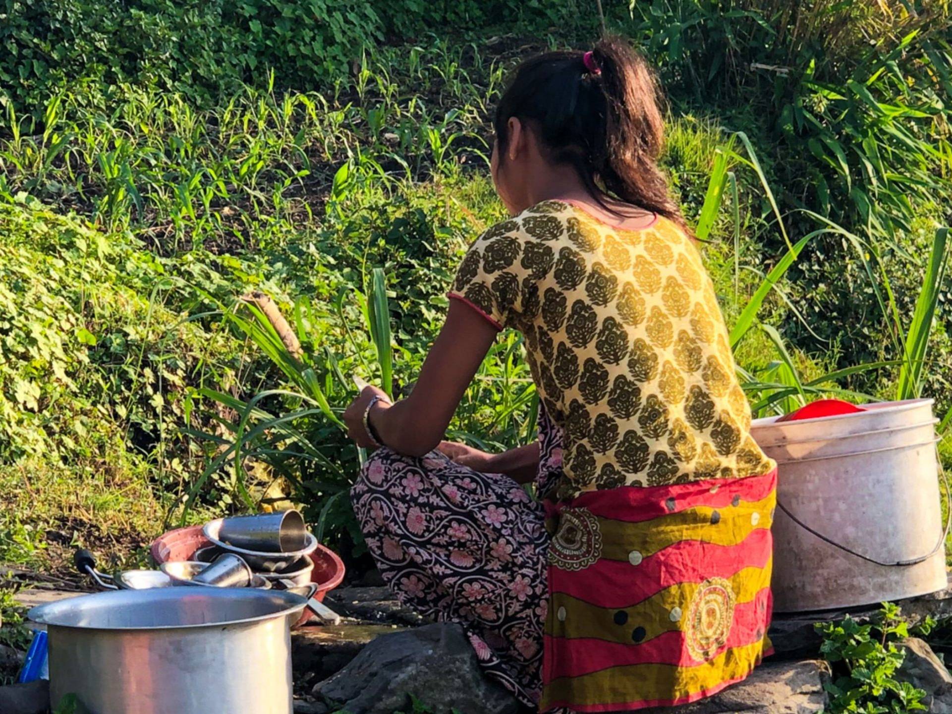 A young woman sits outdoors, surrounded by lush greenery, washing dishes with buckets and utensils by her side, illuminated by the warm sunlight.