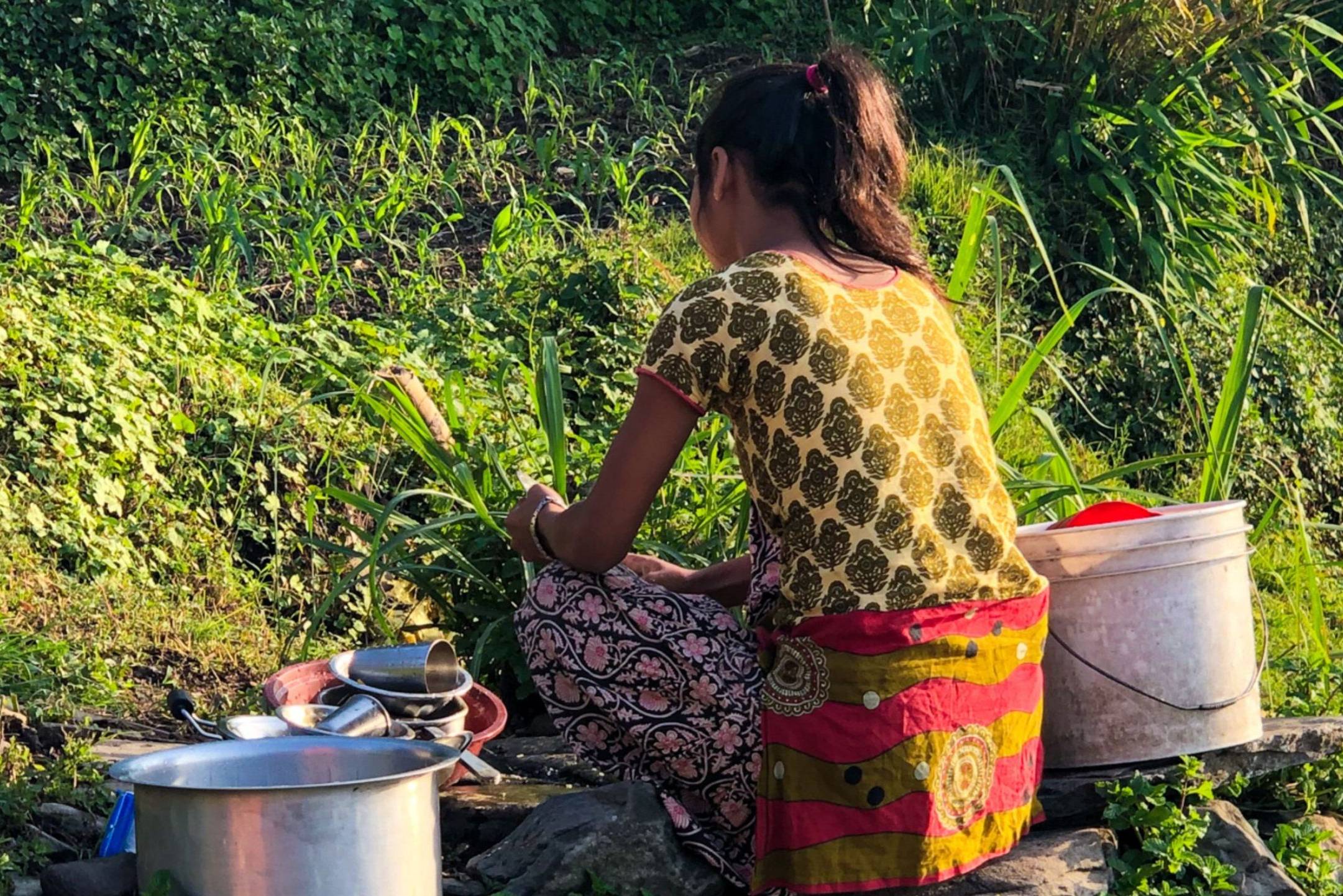 A young woman sits outdoors, surrounded by lush greenery, washing dishes with buckets and utensils by her side, illuminated by the warm sunlight.