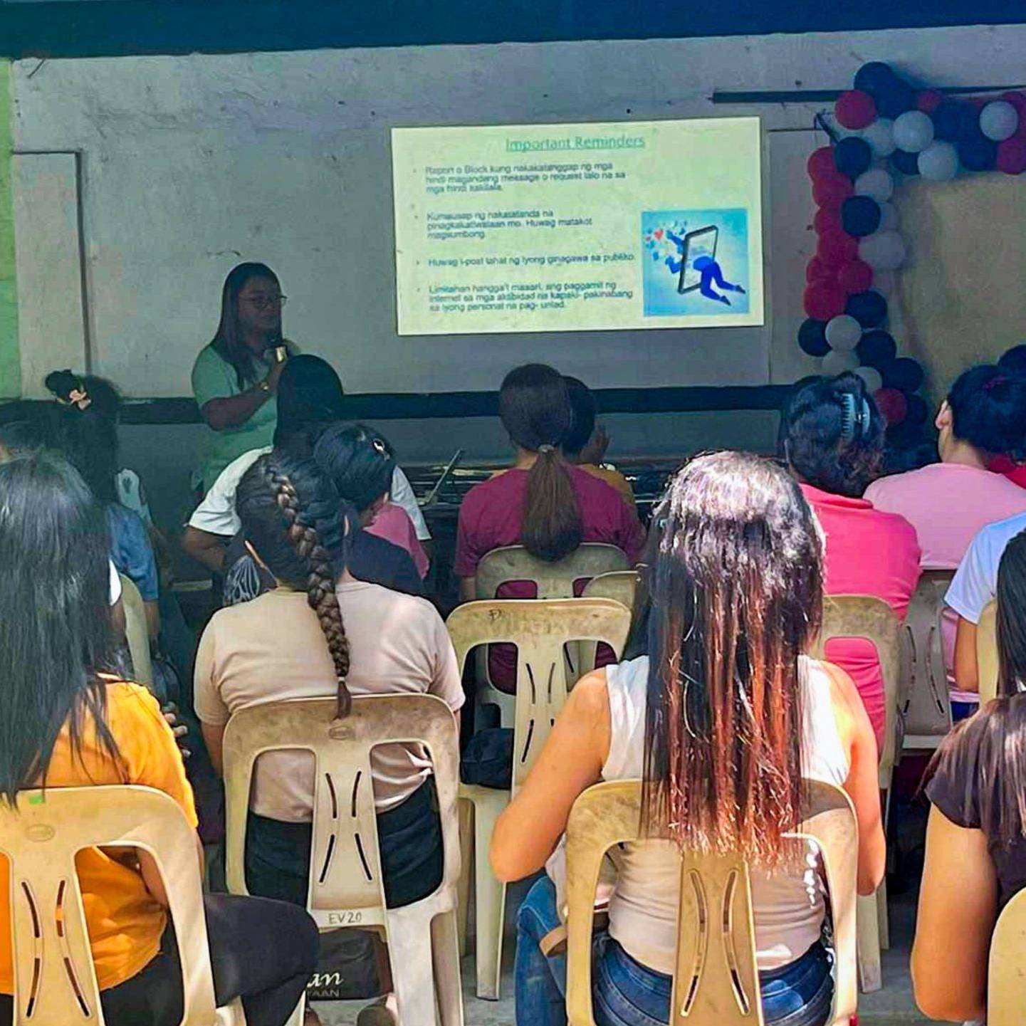 A group of parents attentively listens to a social worker giving an awareness lecture, with a presentation projected in the background.