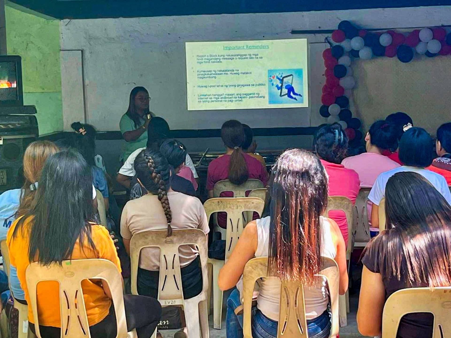 A group of parents attentively listens to a social worker giving an awareness lecture, with a presentation projected in the background.