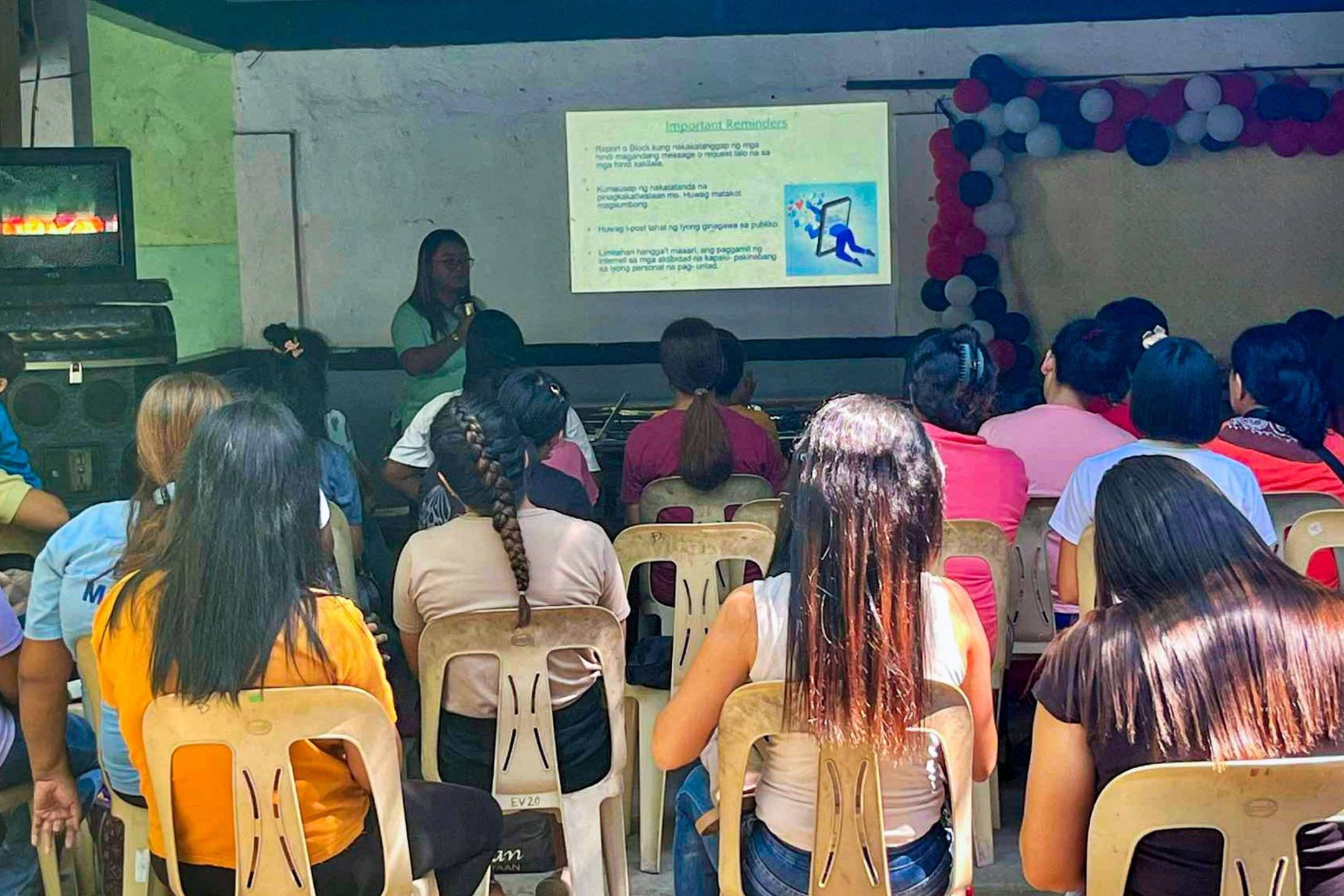 A group of parents attentively listens to a social worker giving an awareness lecture, with a presentation projected in the background.