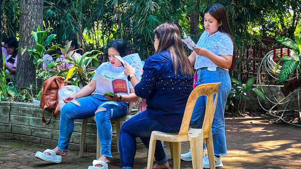 Three women review informational pamphlets outdoors, surrounded by greenery and a relaxed setting.