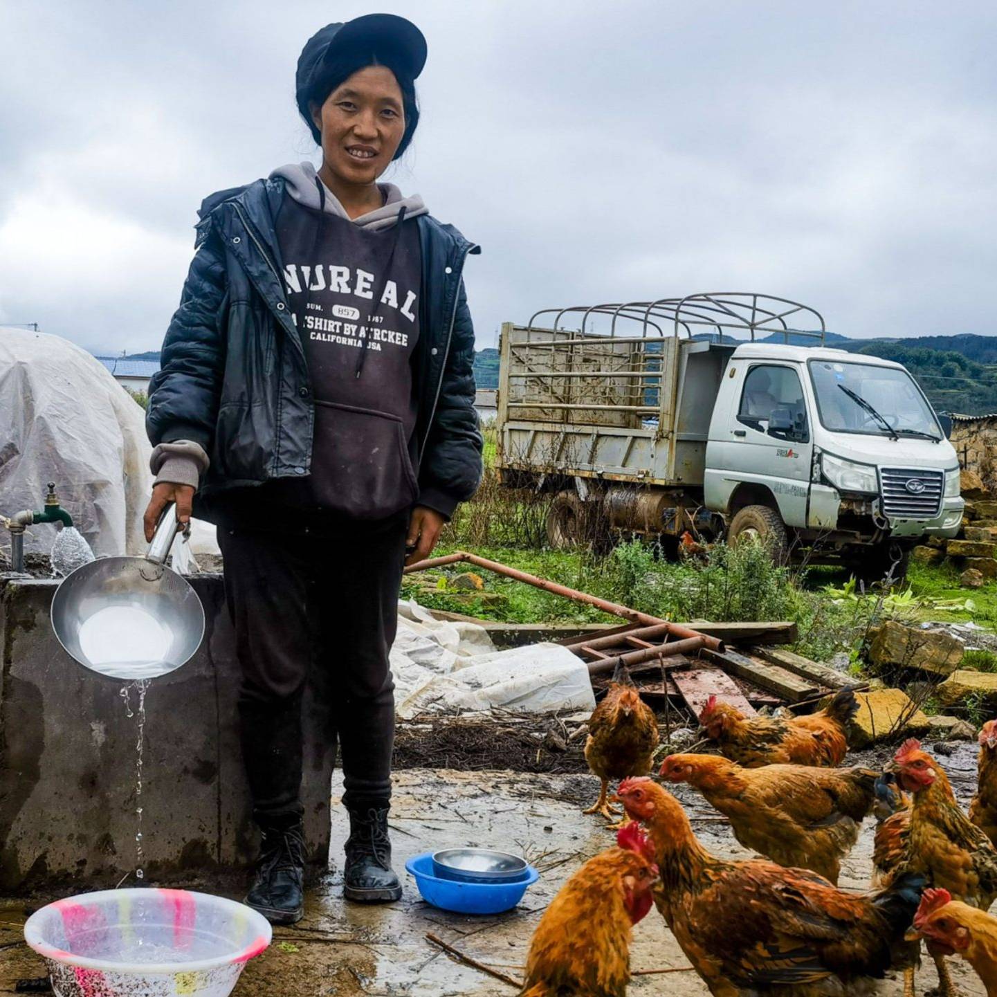 A woman in a black jacket and cap pours water from a pan while surrounded by chickens in a rural farm setting.