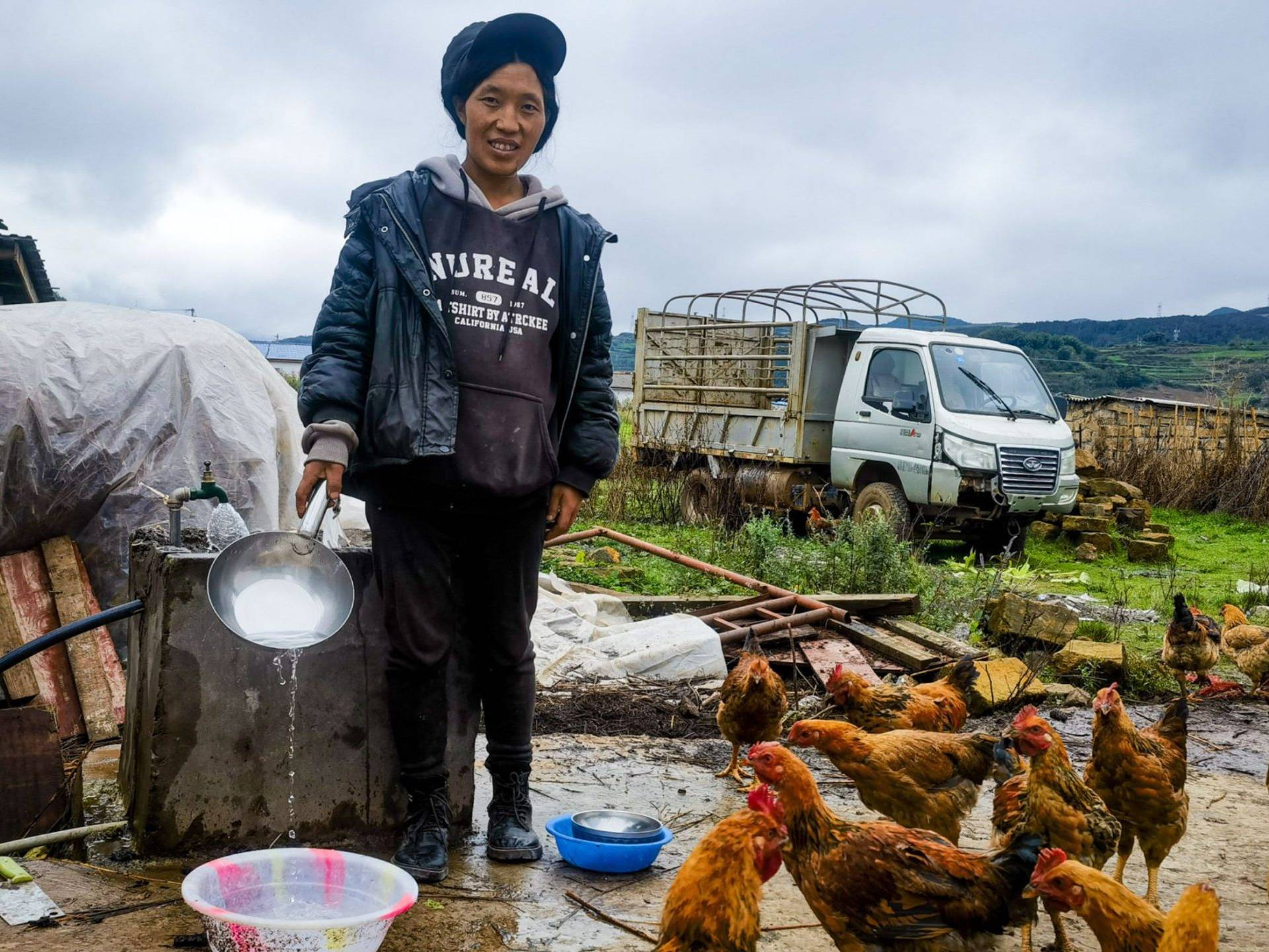 A woman in a black jacket and cap pours water from a pan while surrounded by chickens in a rural farm setting.