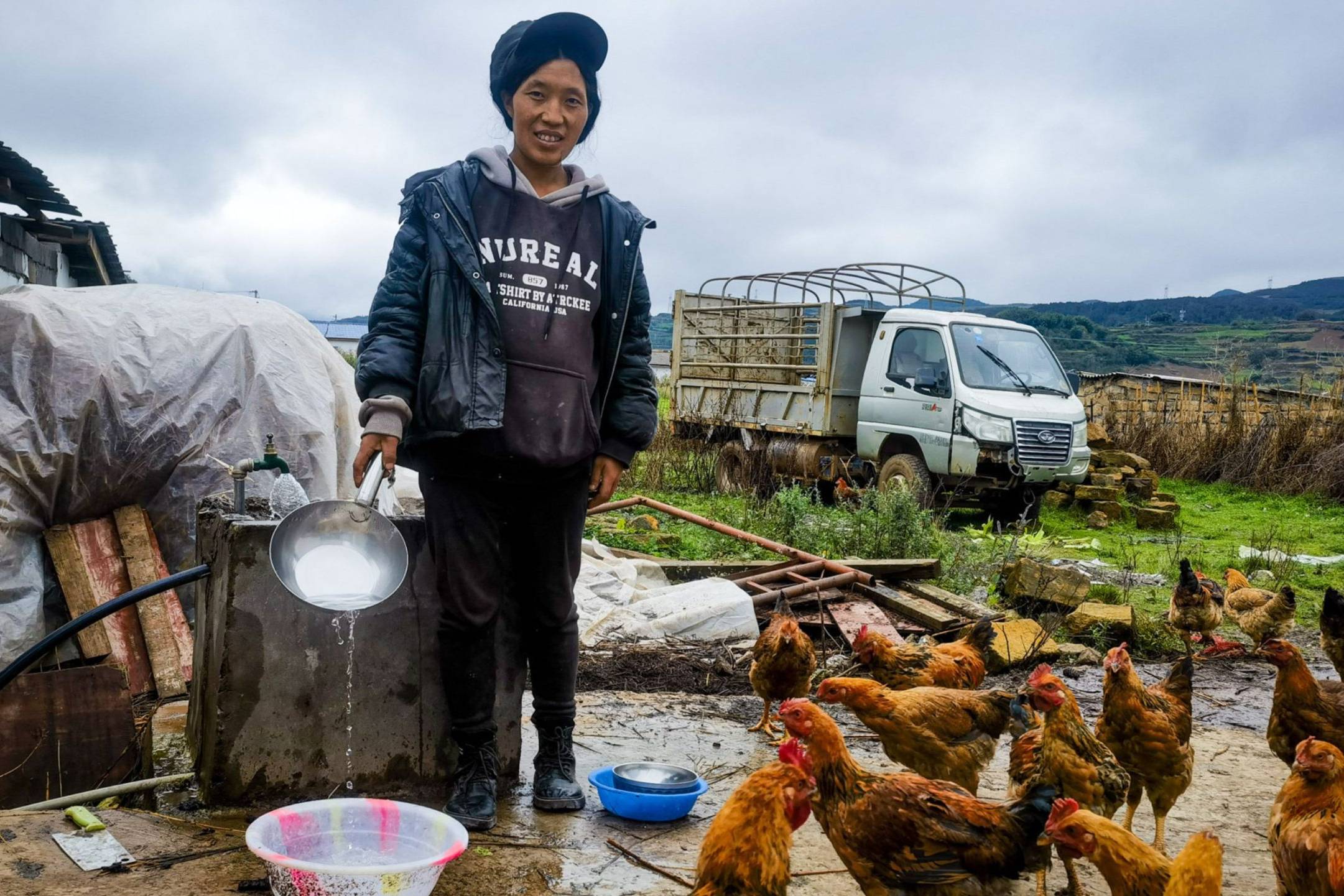 A woman in a black jacket and cap pours water from a pan while surrounded by chickens in a rural farm setting.