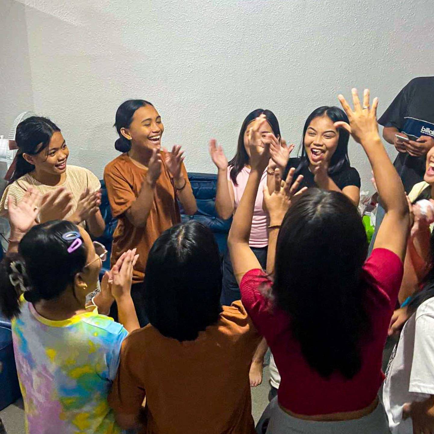 A lively group of young people in a room, all raising their hands enthusiastically in celebration or excitement.