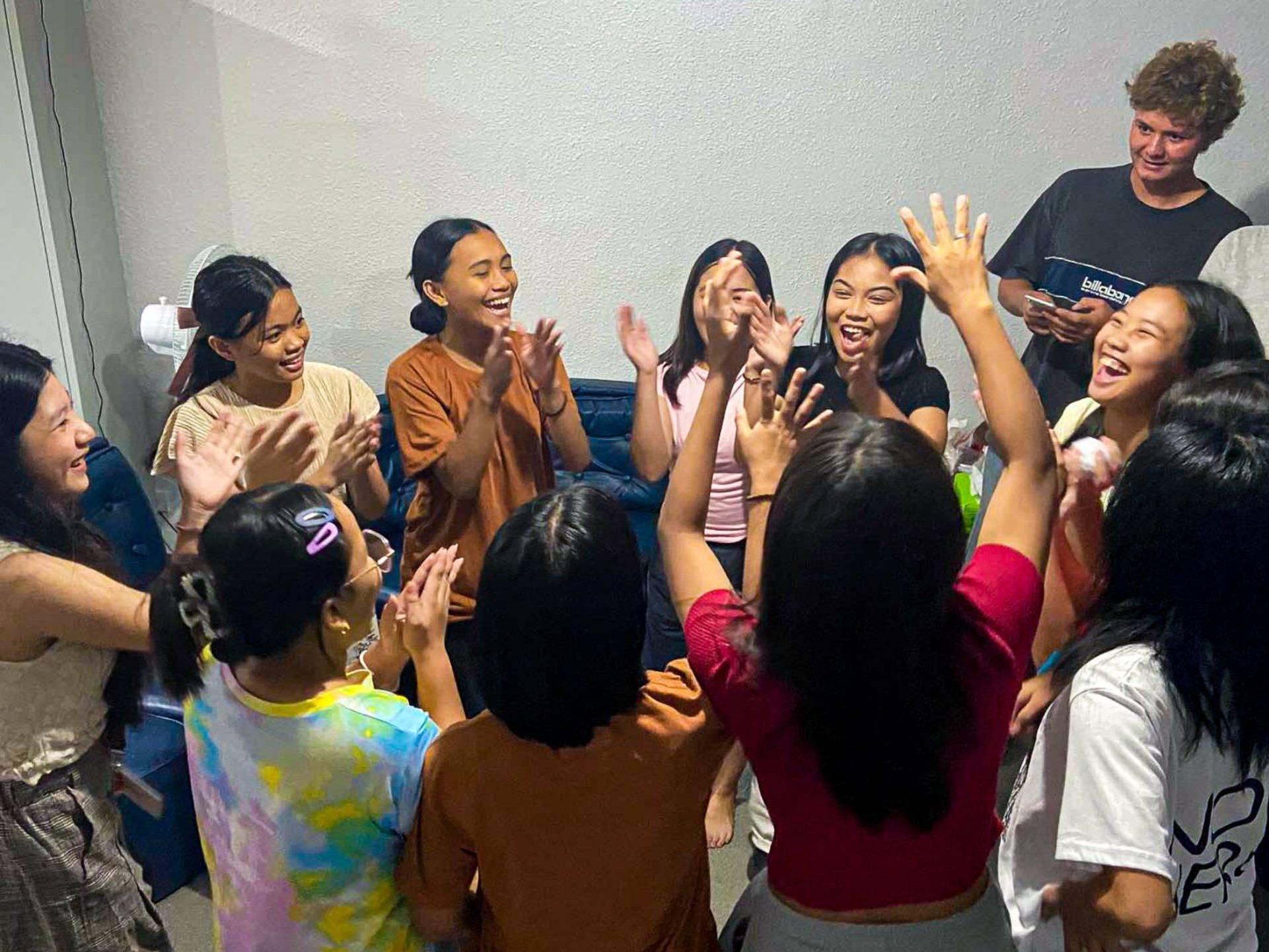 A lively group of young people in a room, all raising their hands enthusiastically in celebration or excitement.