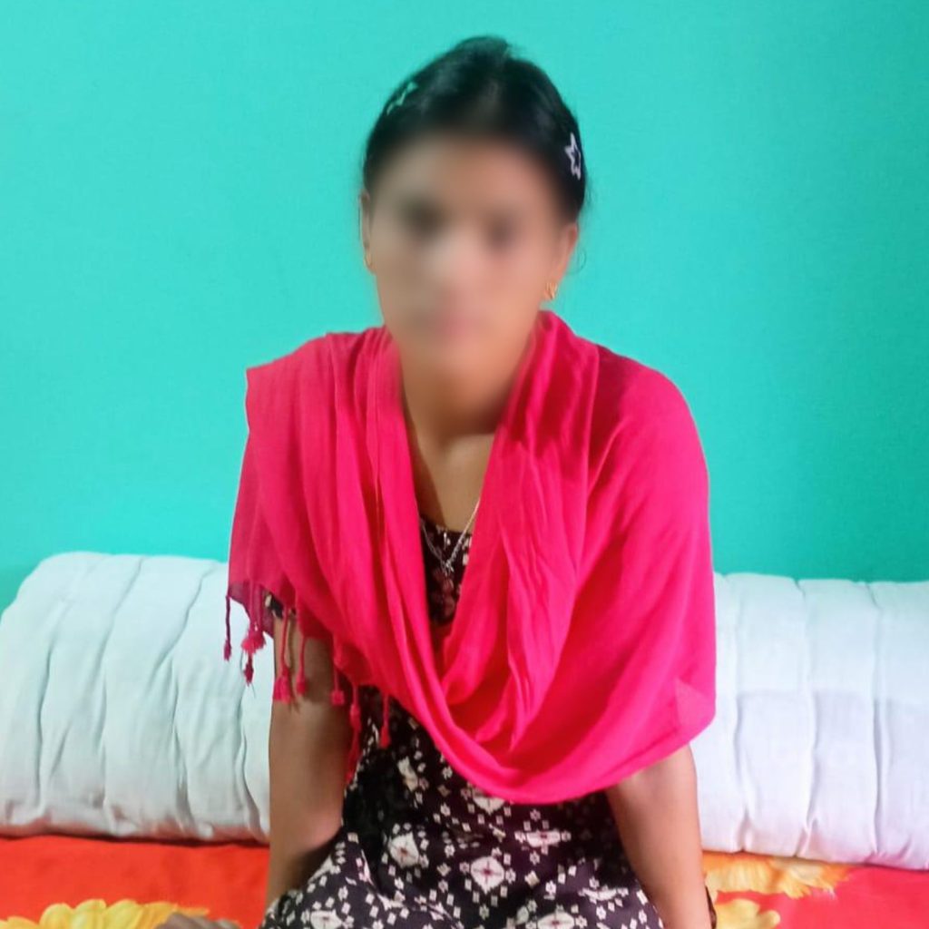 A young woman sits on a bed, wearing a bright pink scarf, her face blurred for privacy.