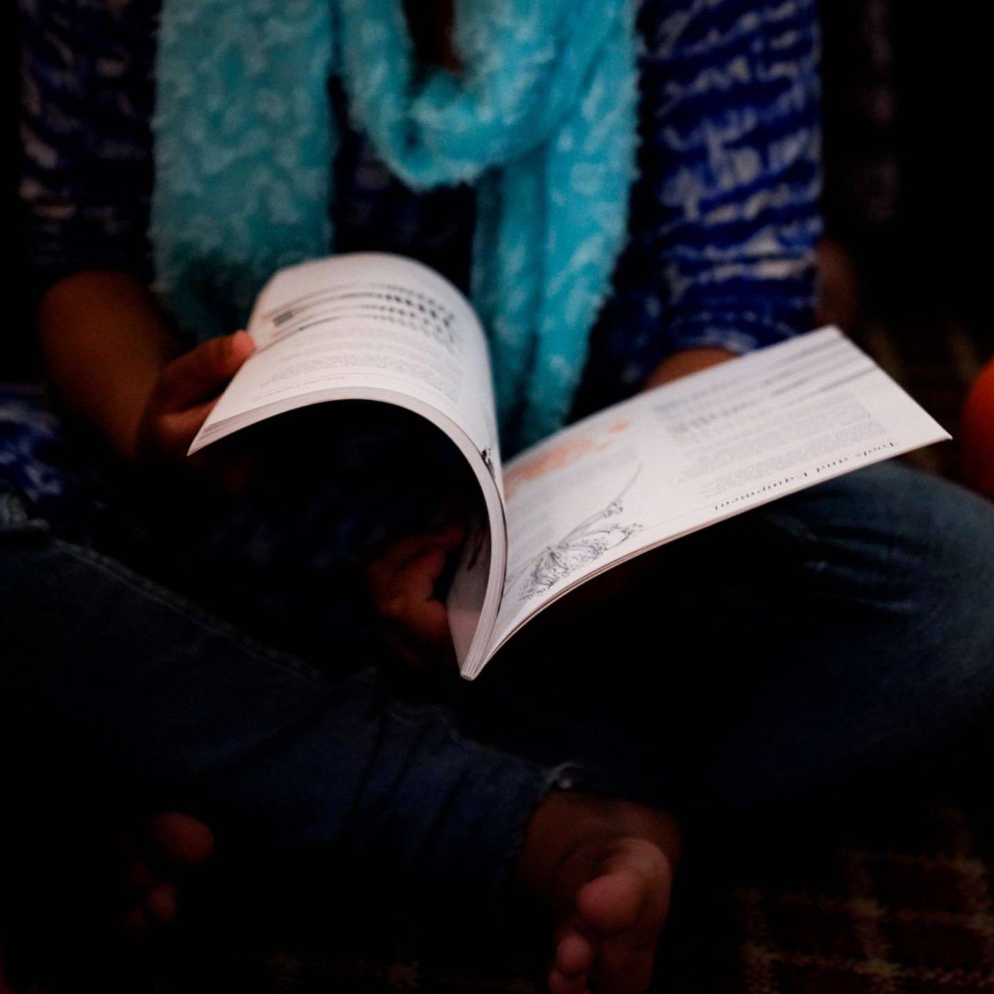 A woman wearing a blue and white outfit and scarf, sitting cross-legged while reading an open book.