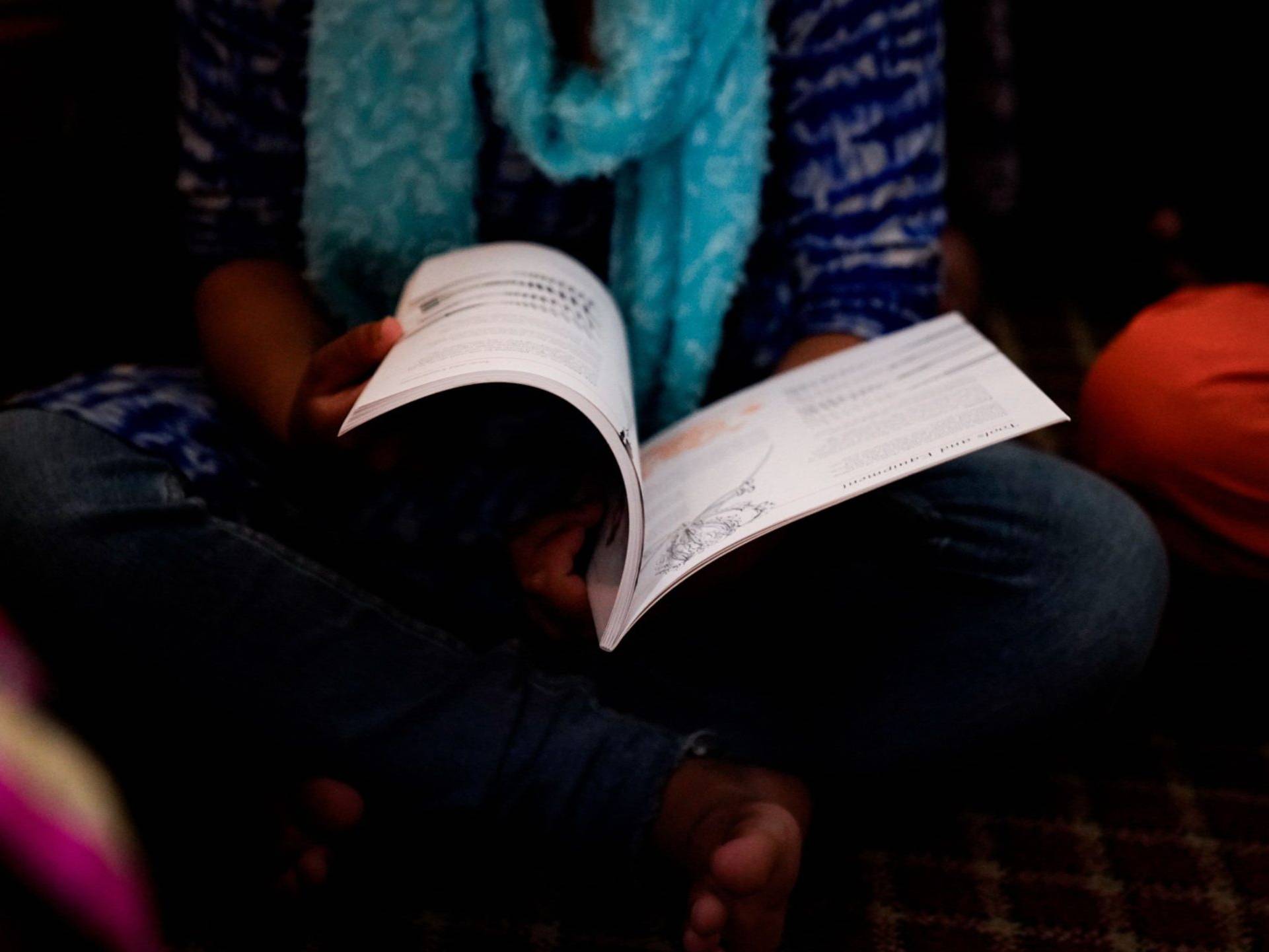 A woman wearing a blue and white outfit and scarf, sitting cross-legged while reading an open book.