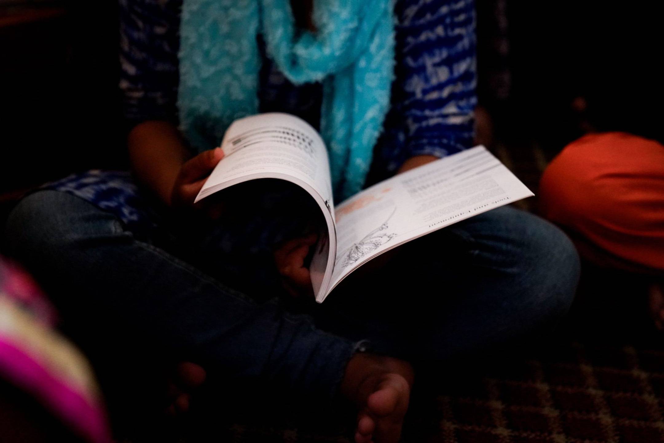 A woman wearing a blue and white outfit and scarf, sitting cross-legged while reading an open book.