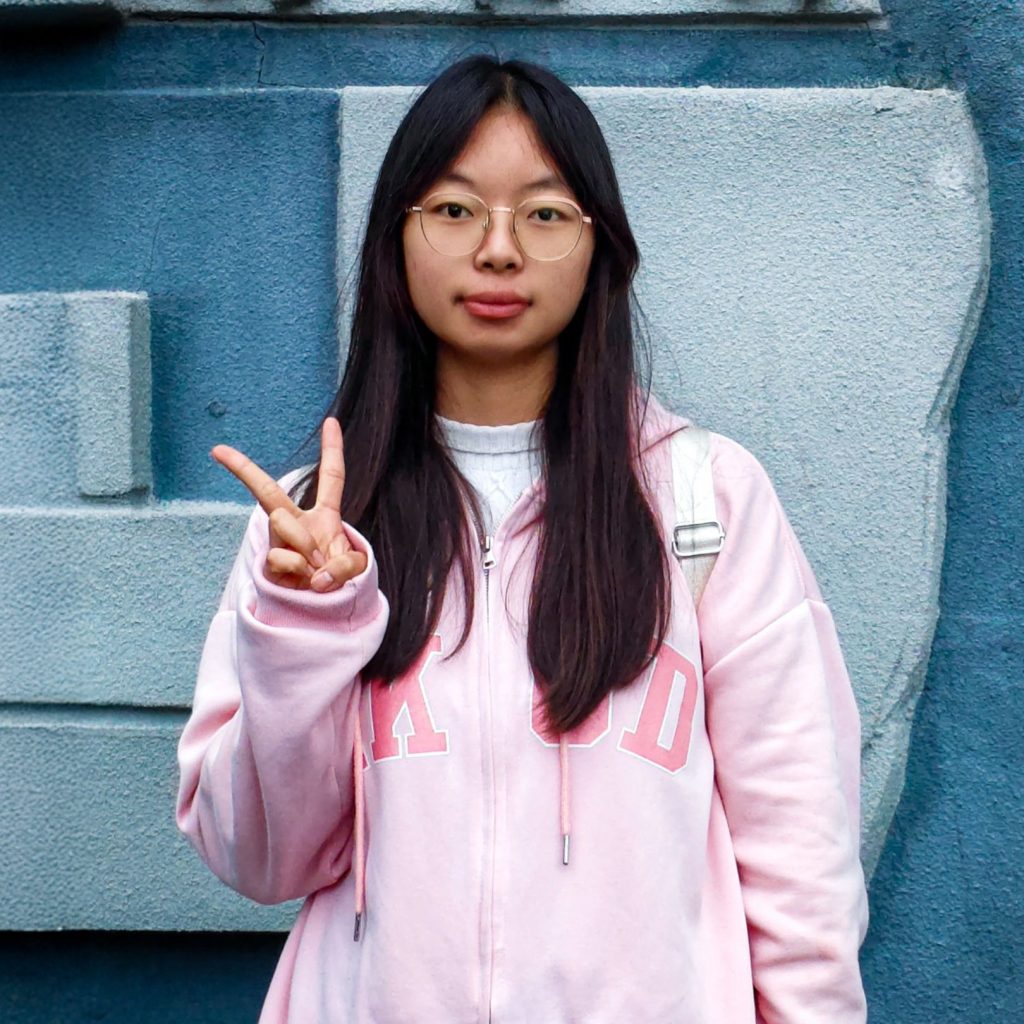 Young woman with long black hair and glasses, wearing a pink and white jacket, standing against a blue wall while making a peace sign.