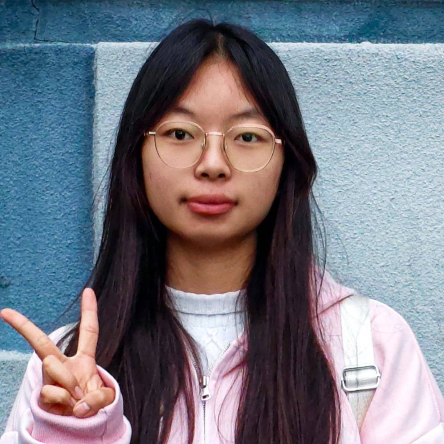 Young woman with long black hair and glasses, wearing a pink and white jacket, standing against a blue wall while making a peace sign.