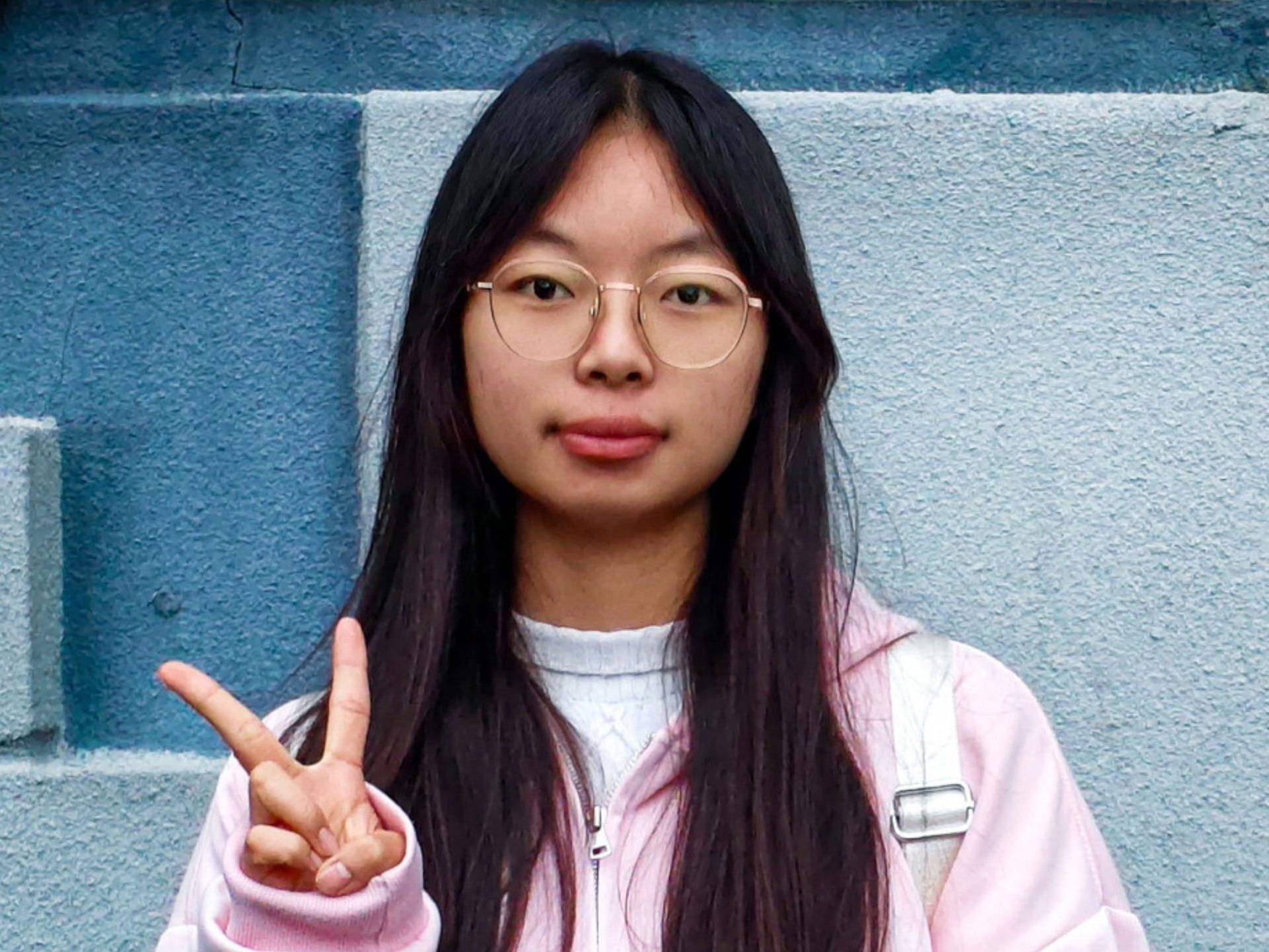 Young woman with long black hair and glasses, wearing a pink and white jacket, standing against a blue wall while making a peace sign.