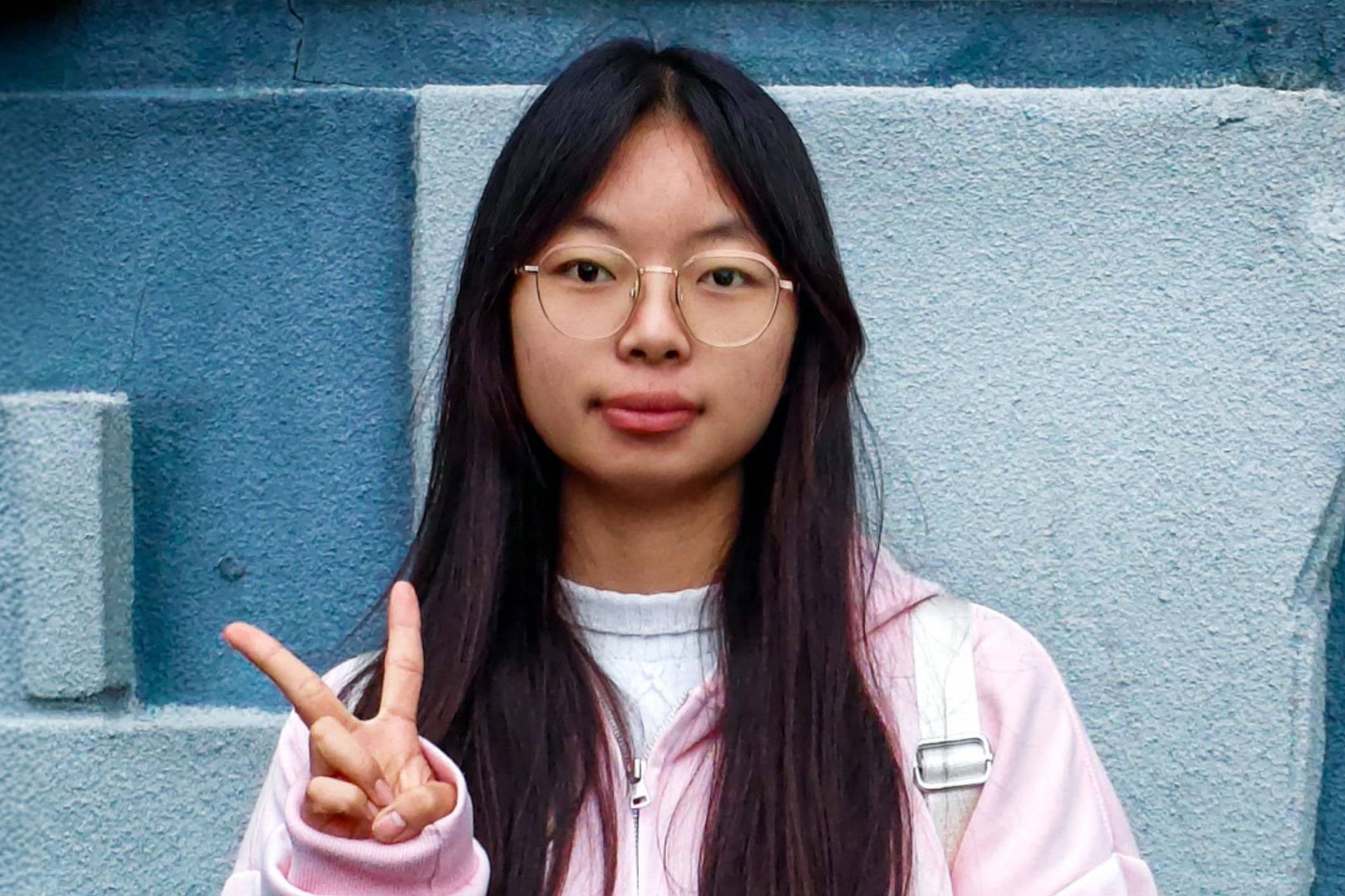 Young woman with long black hair and glasses, wearing a pink and white jacket, standing against a blue wall while making a peace sign.