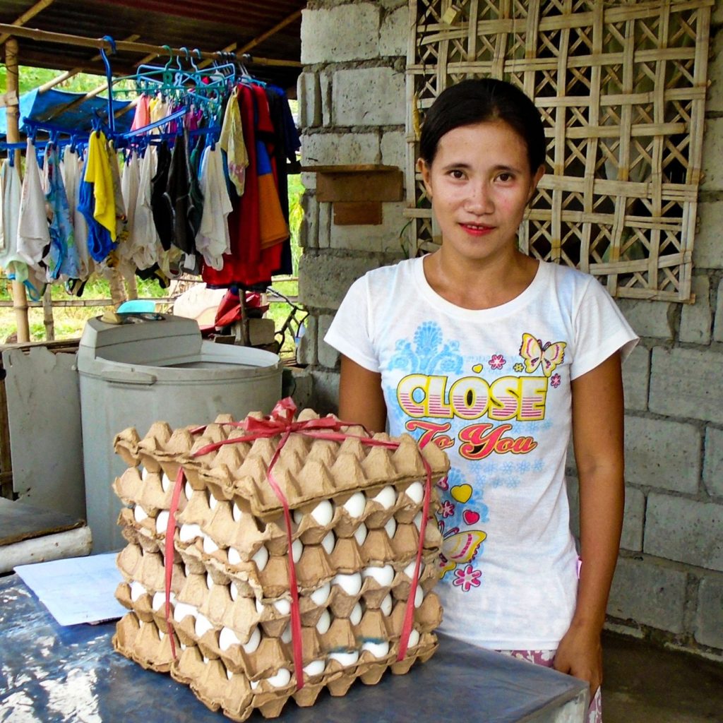 A woman selling eggs poses with neatly stacked trays, surrounded by a home setting with laundry in the background.