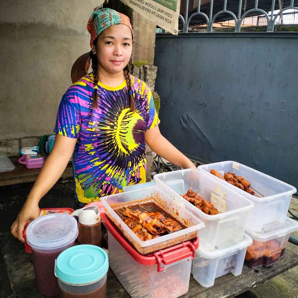 A woman proudly stands behind her food stall, displaying a variety of marinated and cooked dishes.