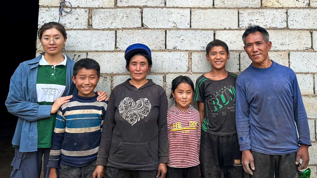 A group of people stands closely together, smiling in front of a rustic brick wall.