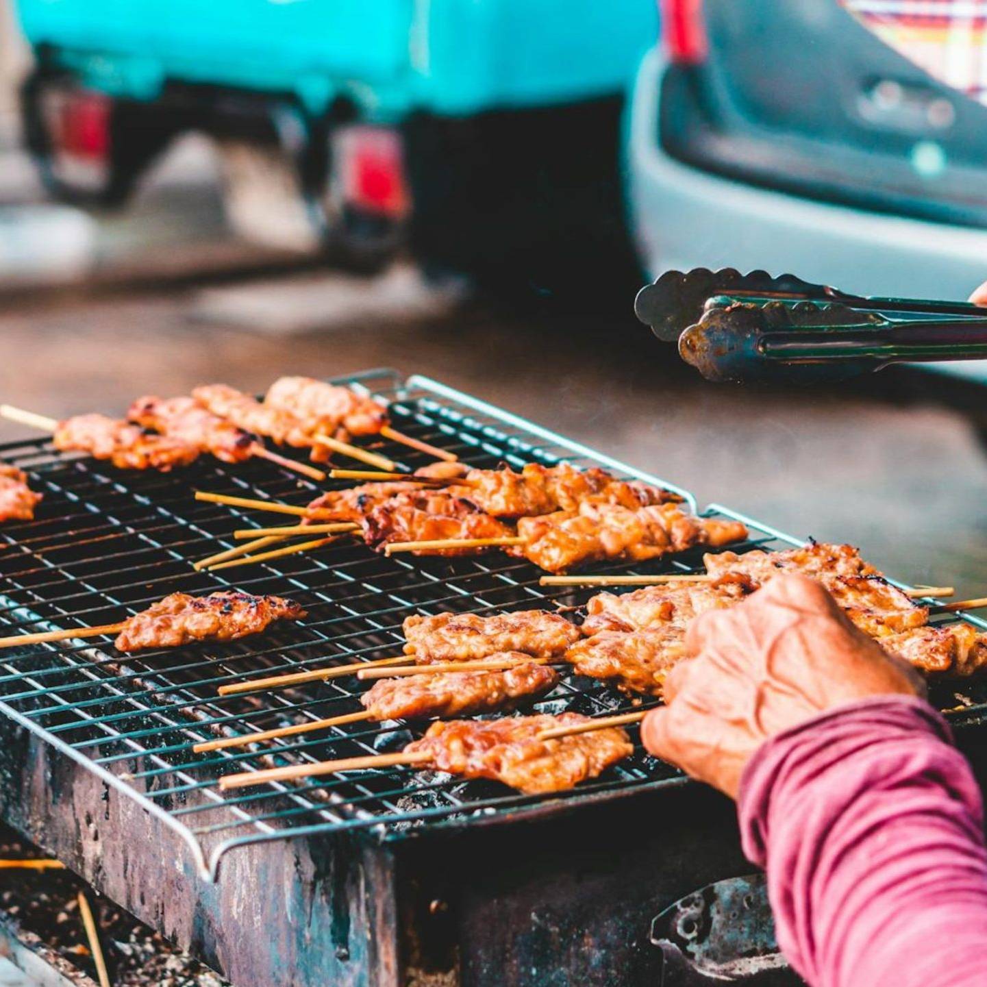 Skewers of meat sizzle on a charcoal grill as a vendor carefully tends to them with tongs.