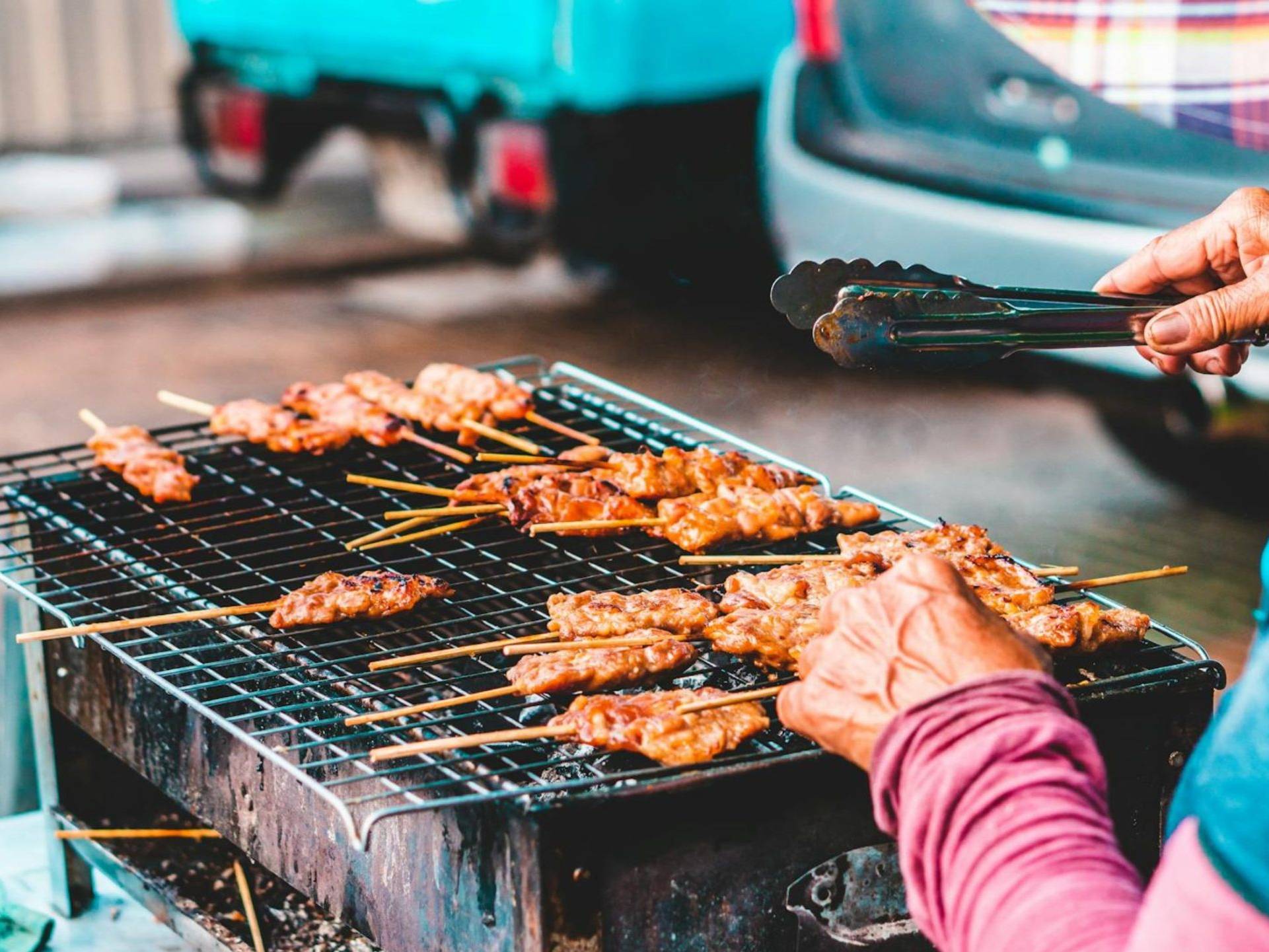 Skewers of meat sizzle on a charcoal grill as a vendor carefully tends to them with tongs.