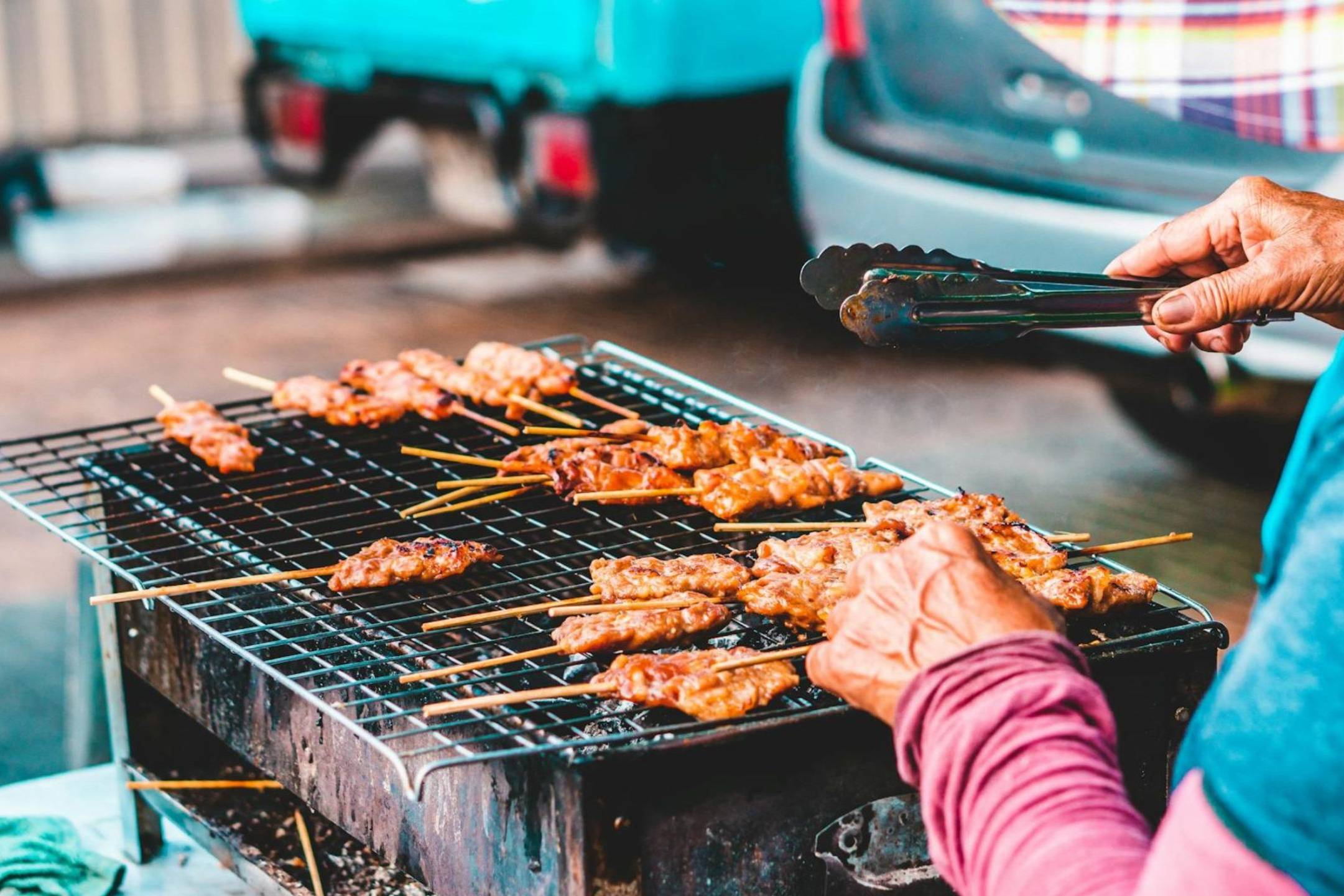 Skewers of meat sizzle on a charcoal grill as a vendor carefully tends to them with tongs.