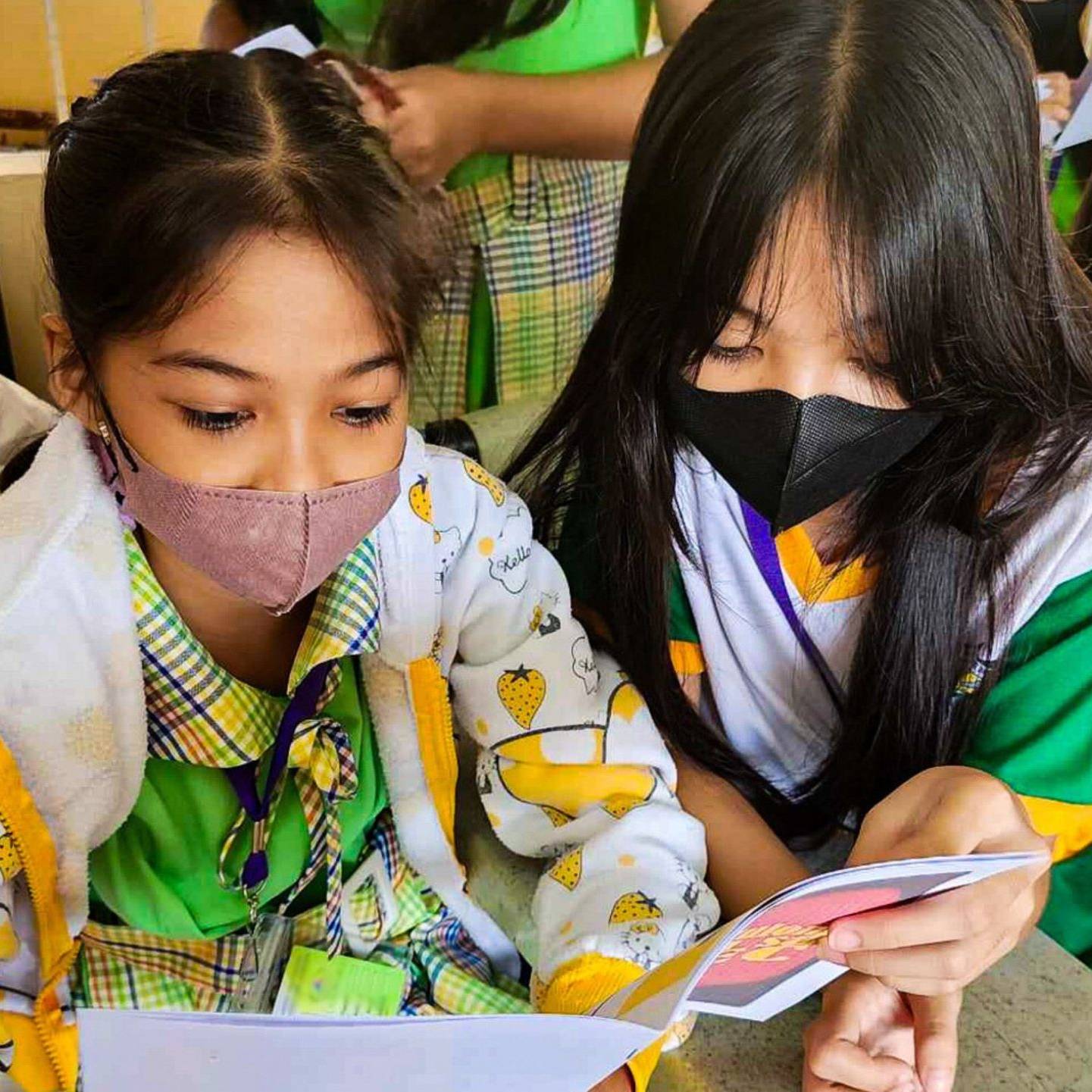 Two young students wearing masks attentively read a booklet together in a classroom.