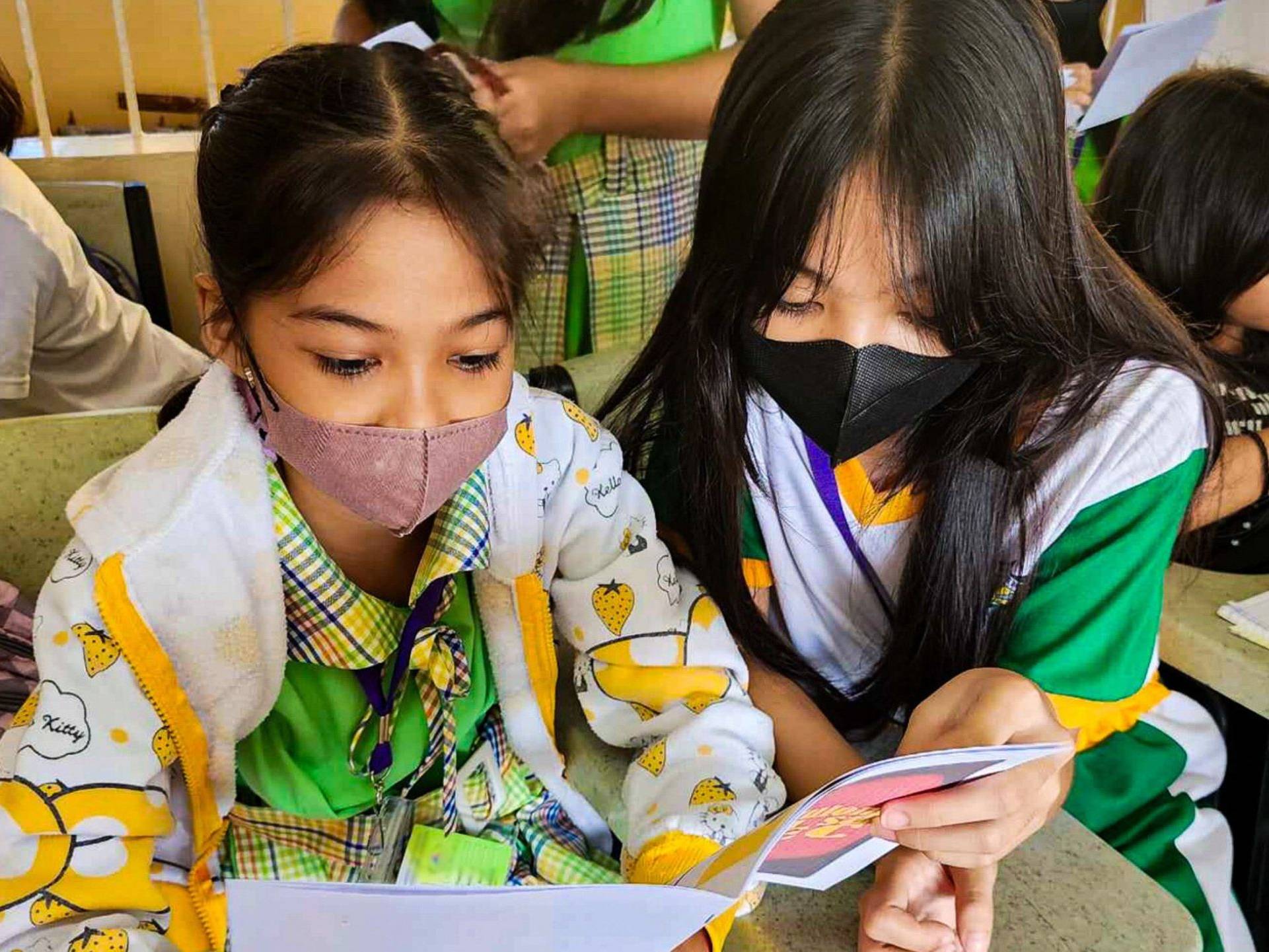 Two young students wearing masks attentively read a booklet together in a classroom.