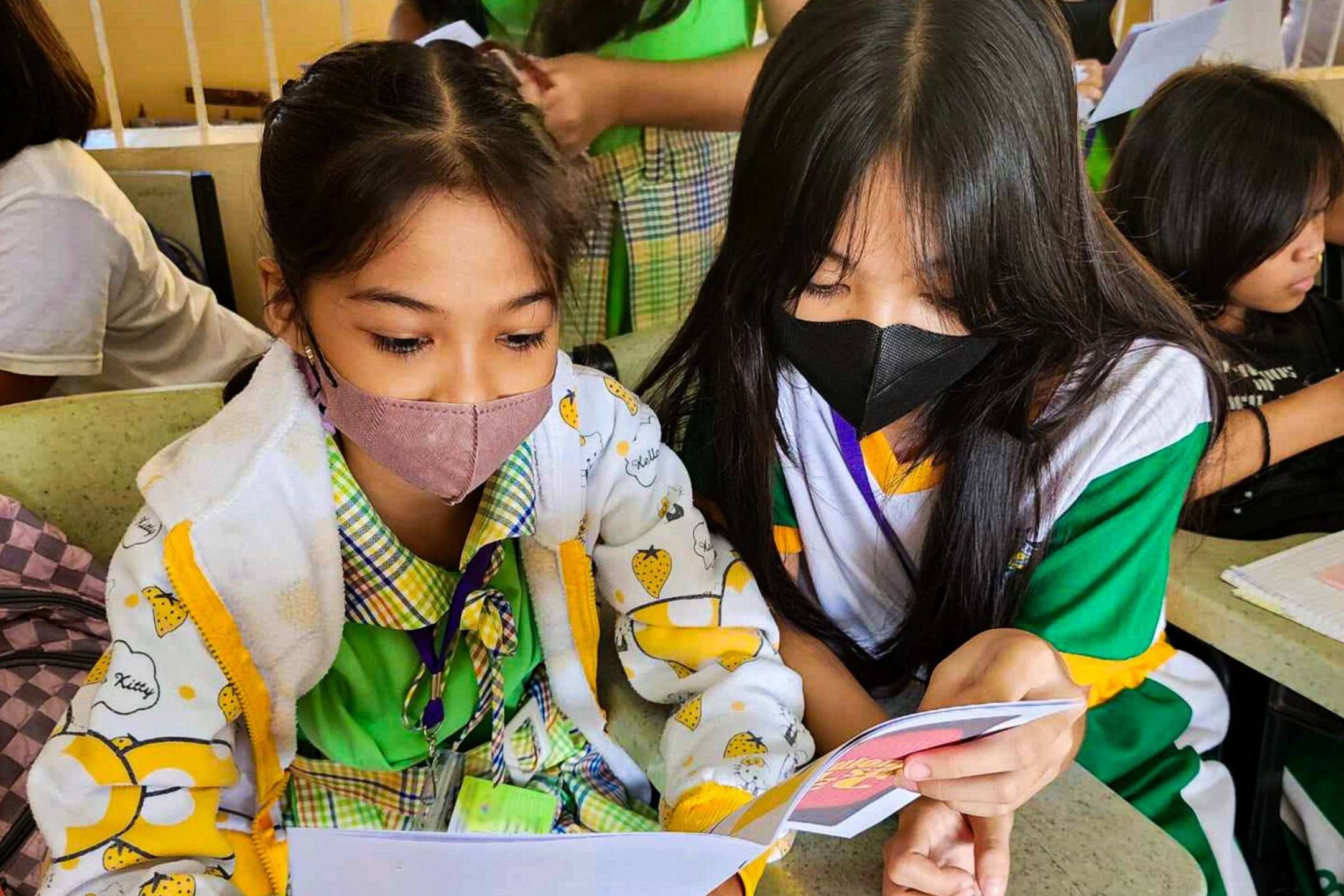 Two young students wearing masks attentively read a booklet together in a classroom.