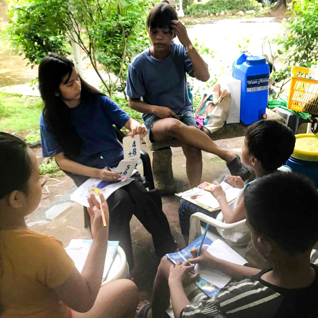 A young woman tutors children outdoors, using flashcards and books.