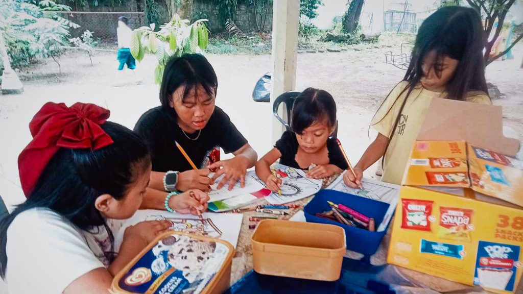 A teenage girl helps young children with coloring and writing activities at an outdoor table.