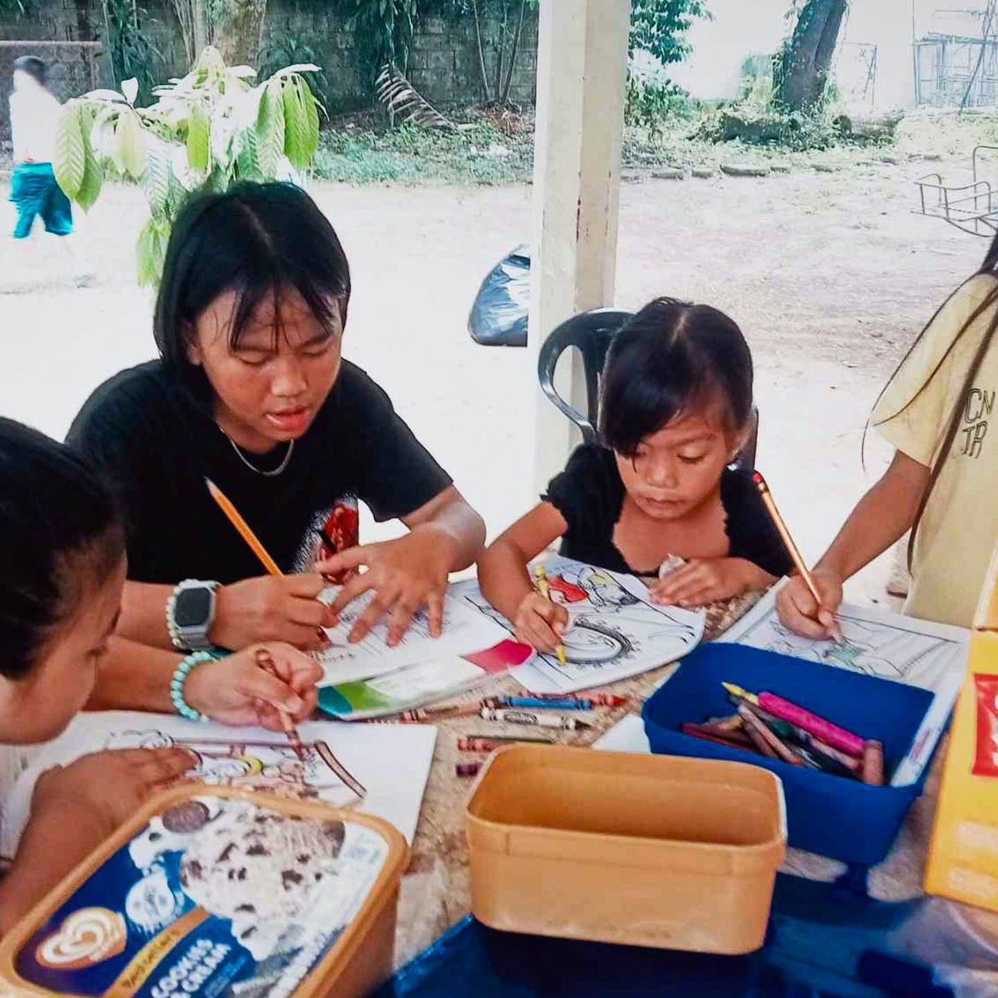 A teenage girl helps young children with coloring and writing activities at an outdoor table.