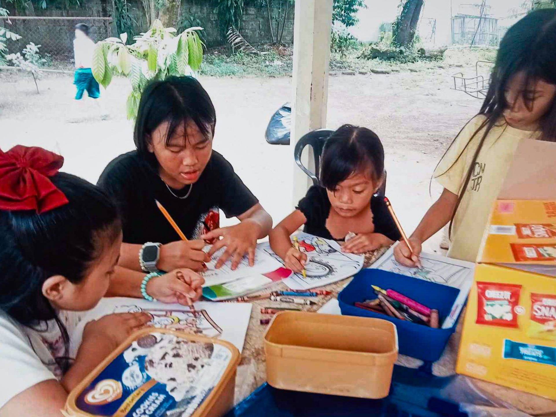 A teenage girl helps young children with coloring and writing activities at an outdoor table.