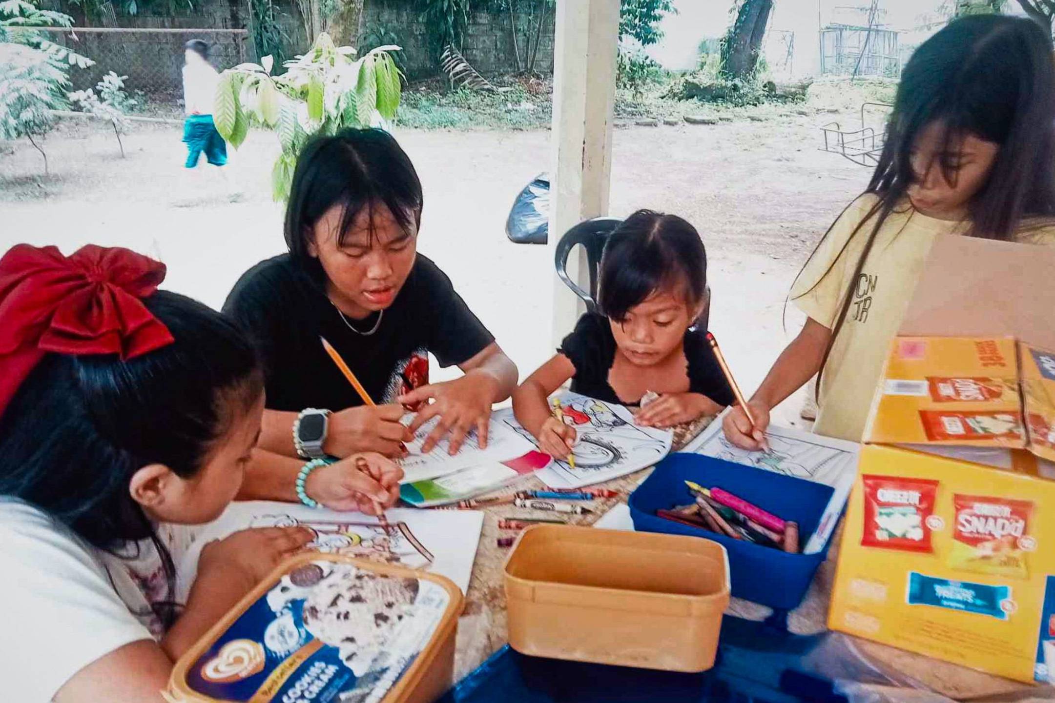 A teenage girl helps young children with coloring and writing activities at an outdoor table.