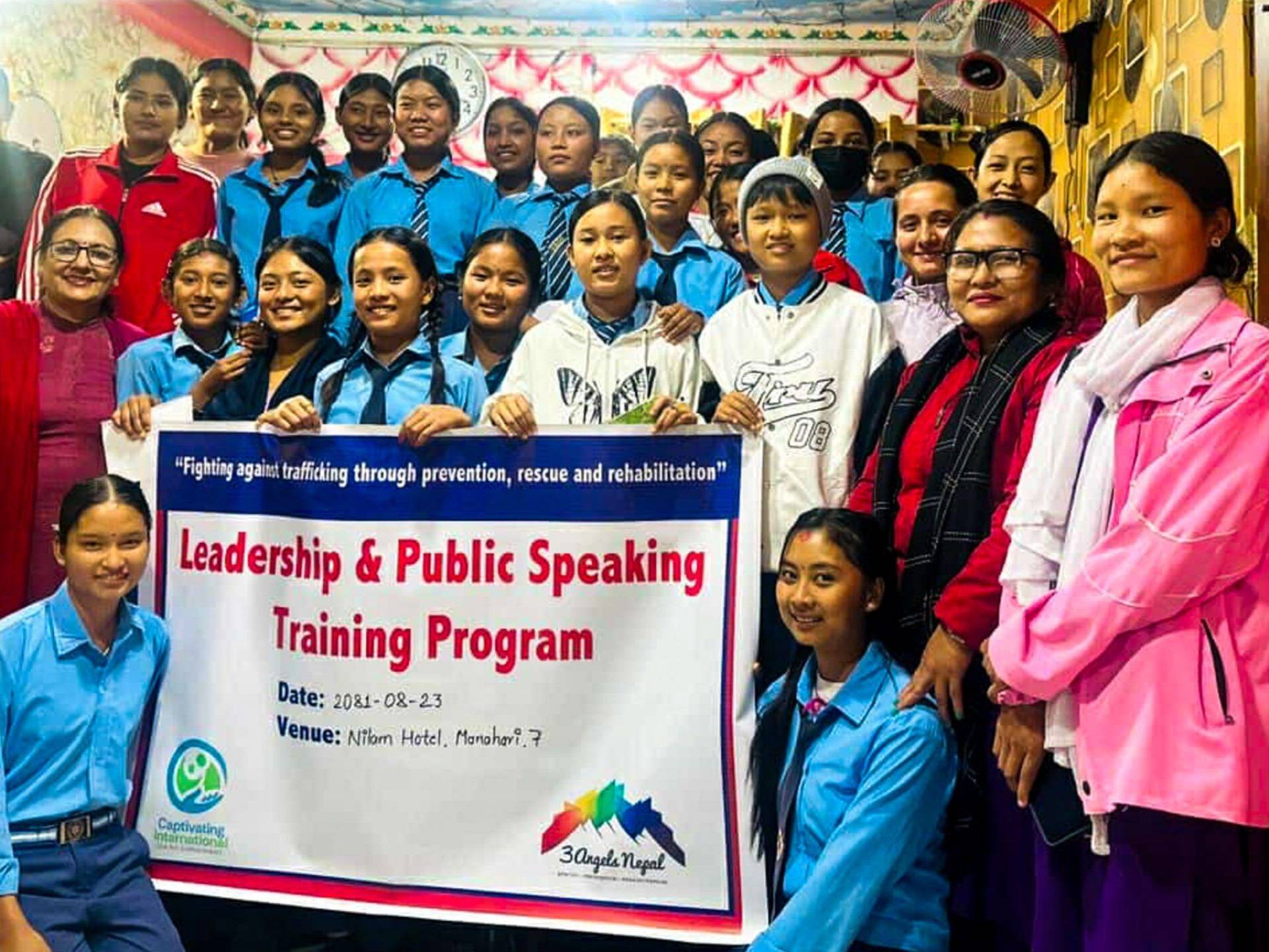 A group of girls and trainers posing with a banner for the Leadership & Public Speaking Training Program.