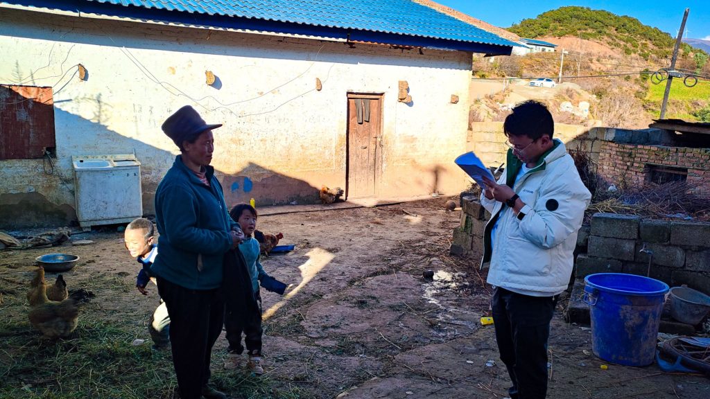 A man interviews a woman outside a house while her two children are playing nearby.