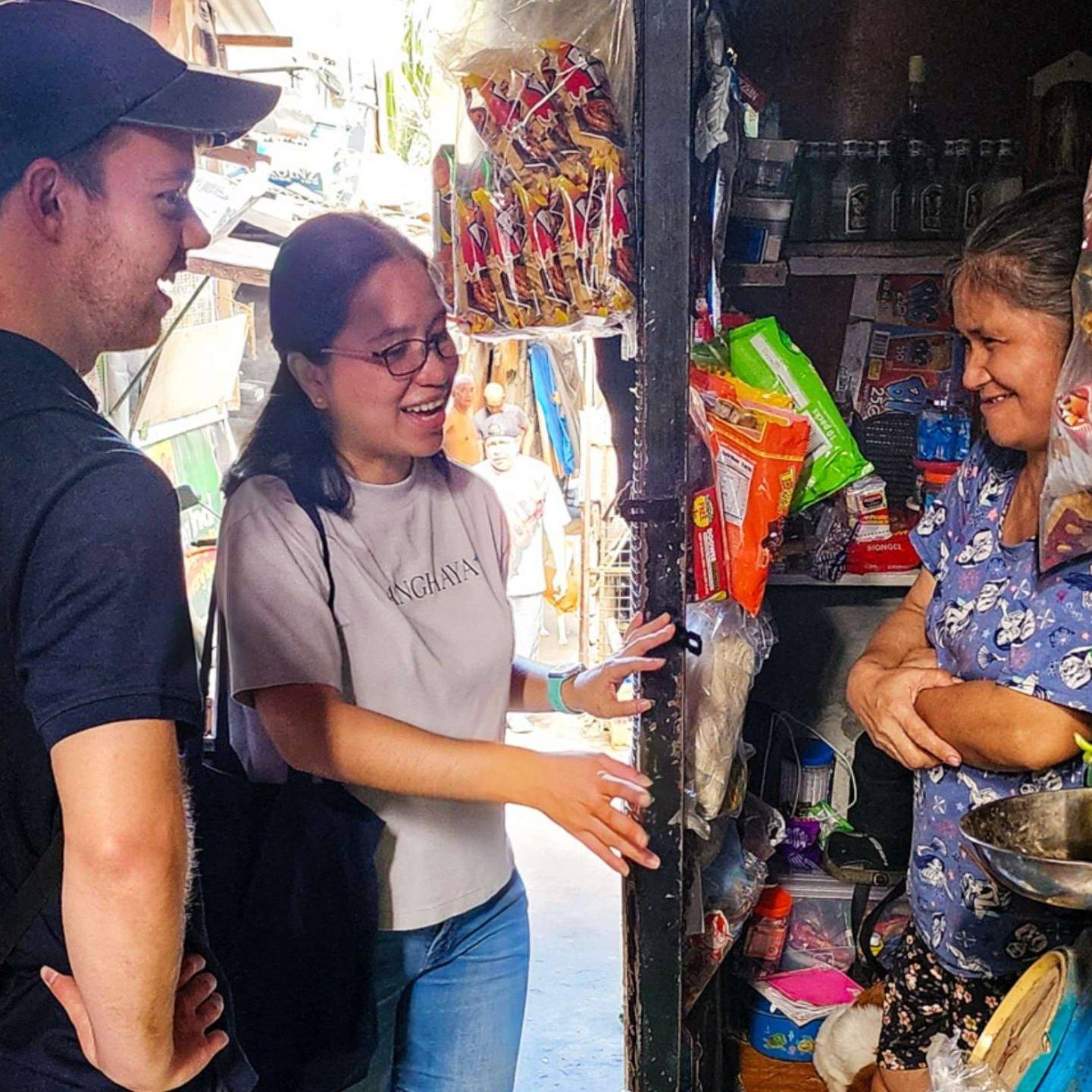 Three people stand outside a food store, smiling, with various food items displayed.