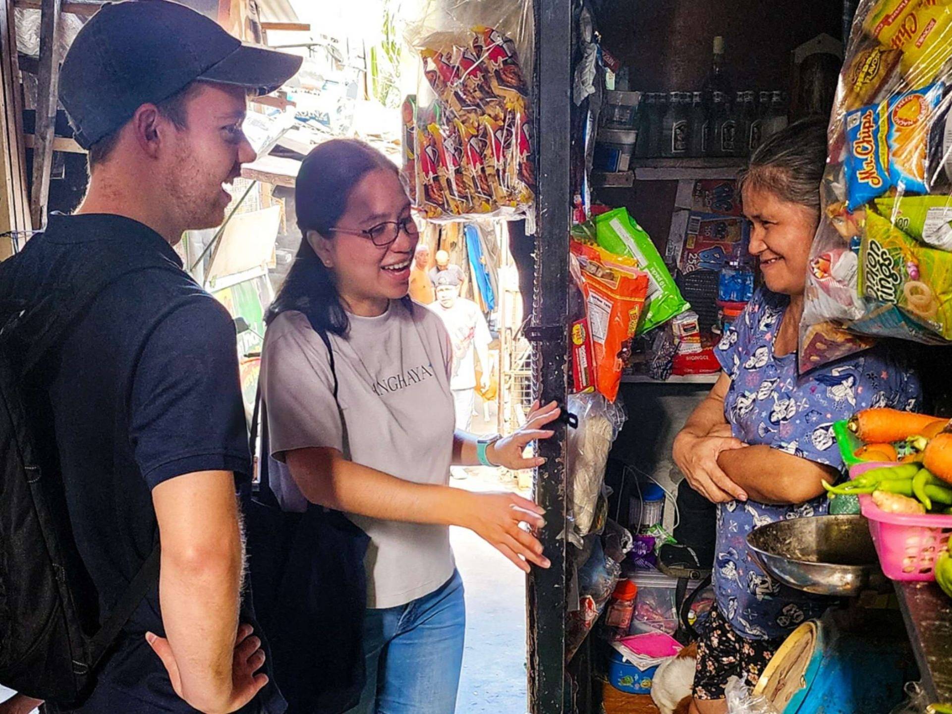 Three people stand outside a food store, smiling, with various food items displayed.