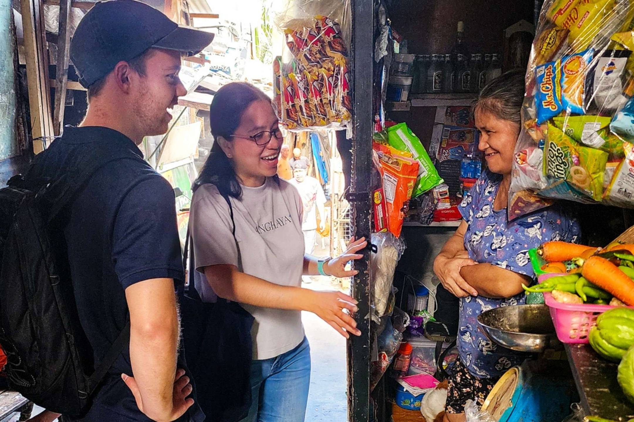 Three people stand outside a food store, smiling, with various food items displayed.