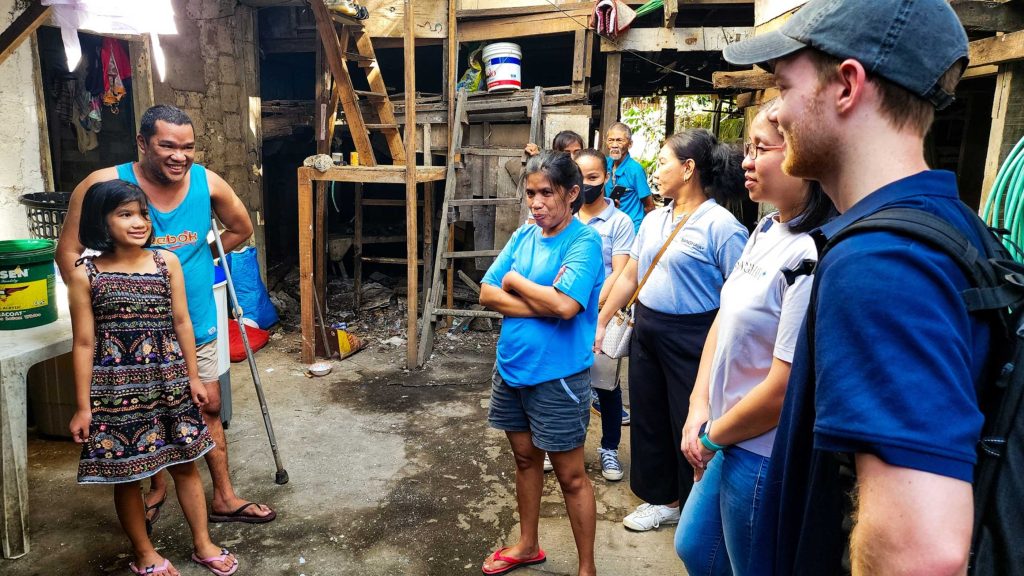 A small group of individuals stands outside a house in a slum area, engaged in conversation amidst their surroundings.