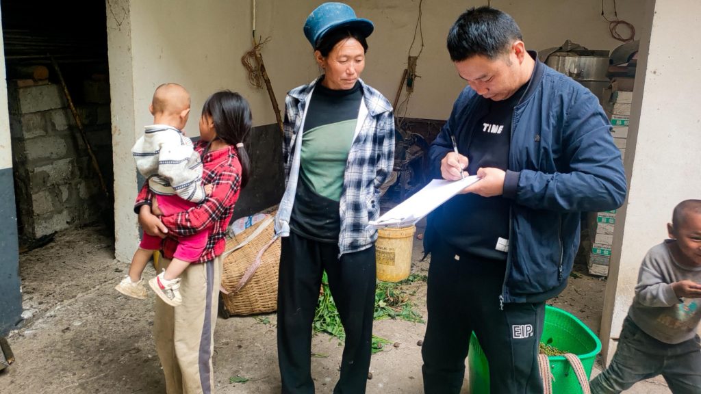 A man writing something down, a woman looking at the paper, and children stand in front of a house.