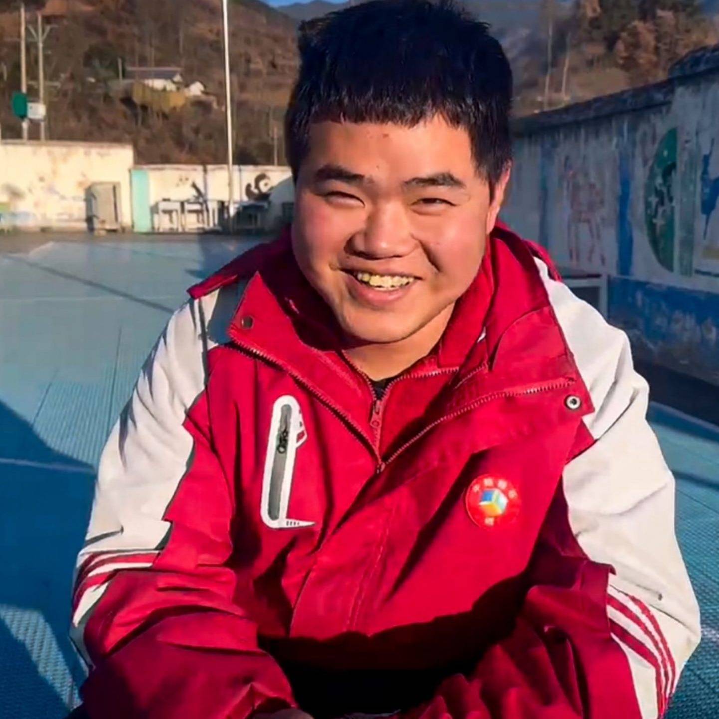 A young man smiling and wearing a red jacket is seated on a basketball court.