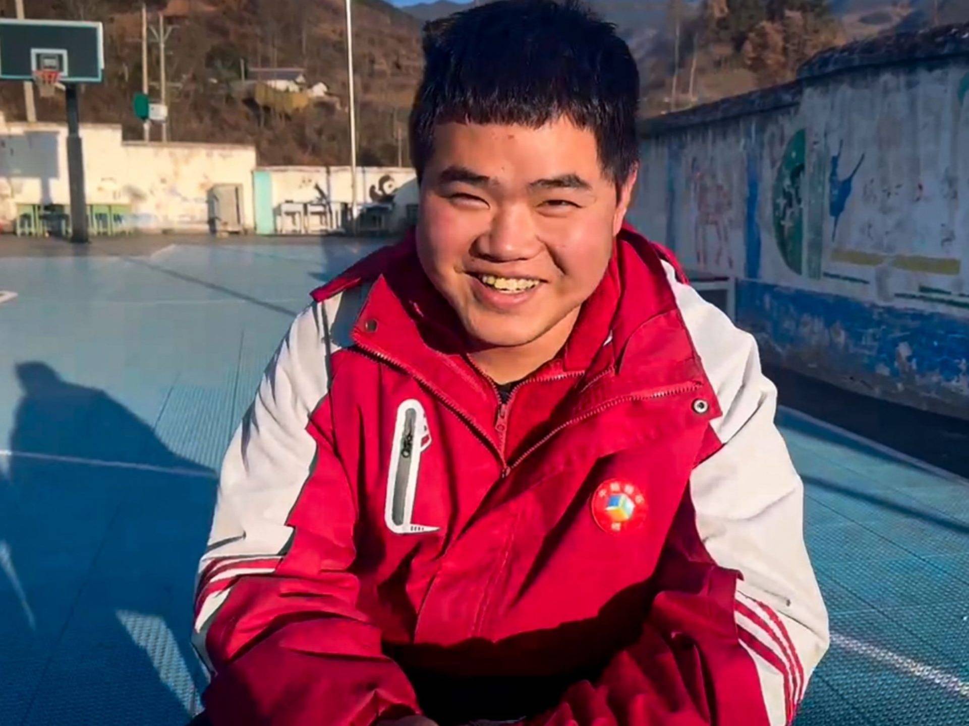 A young man smiling and wearing a red jacket is seated on a basketball court.