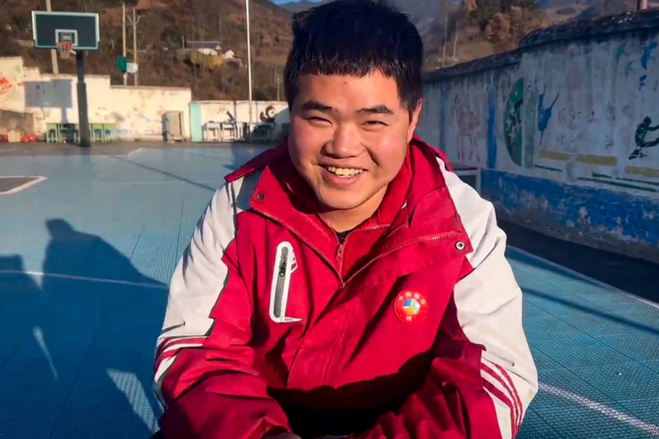 A young man smiling and wearing a red jacket is seated on a basketball court.