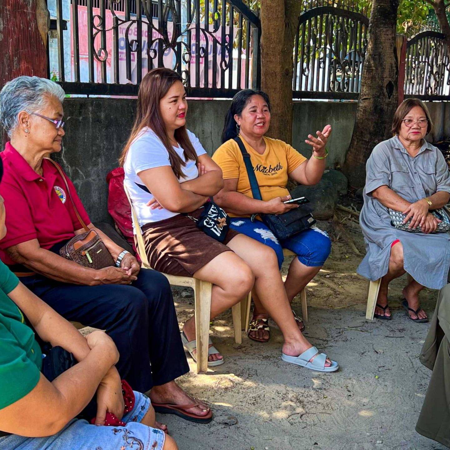 A group of women seated on chairs in front of a fence, engaged in conversation and enjoying their time together.