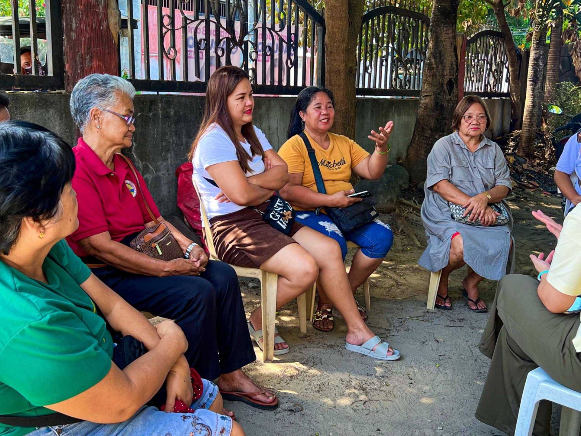 A group of women seated on chairs in front of a fence, engaged in conversation and enjoying their time together.