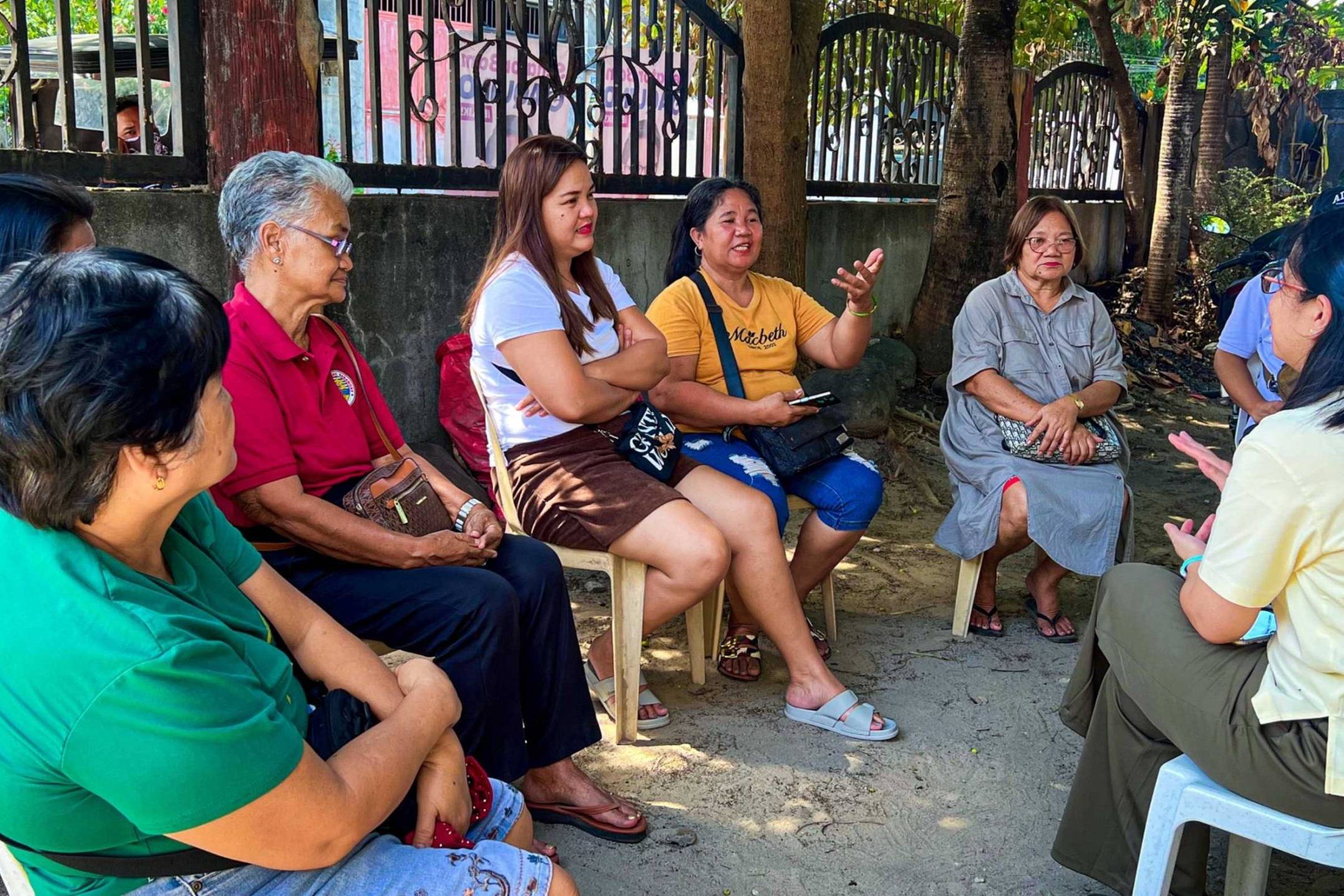 A group of women seated on chairs in front of a fence, engaged in conversation and enjoying their time together.