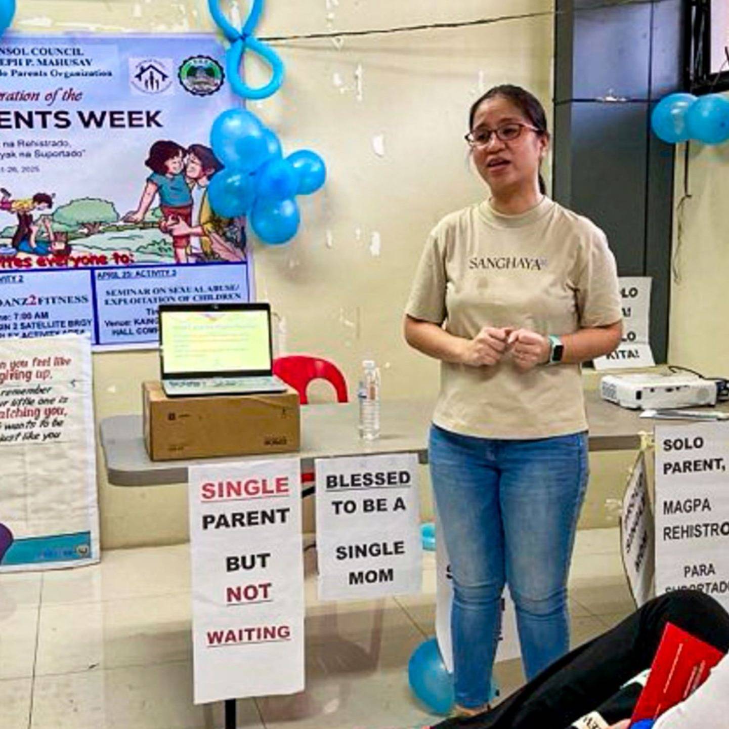 A woman poses in front of a table speaking in front of an audience in a room.