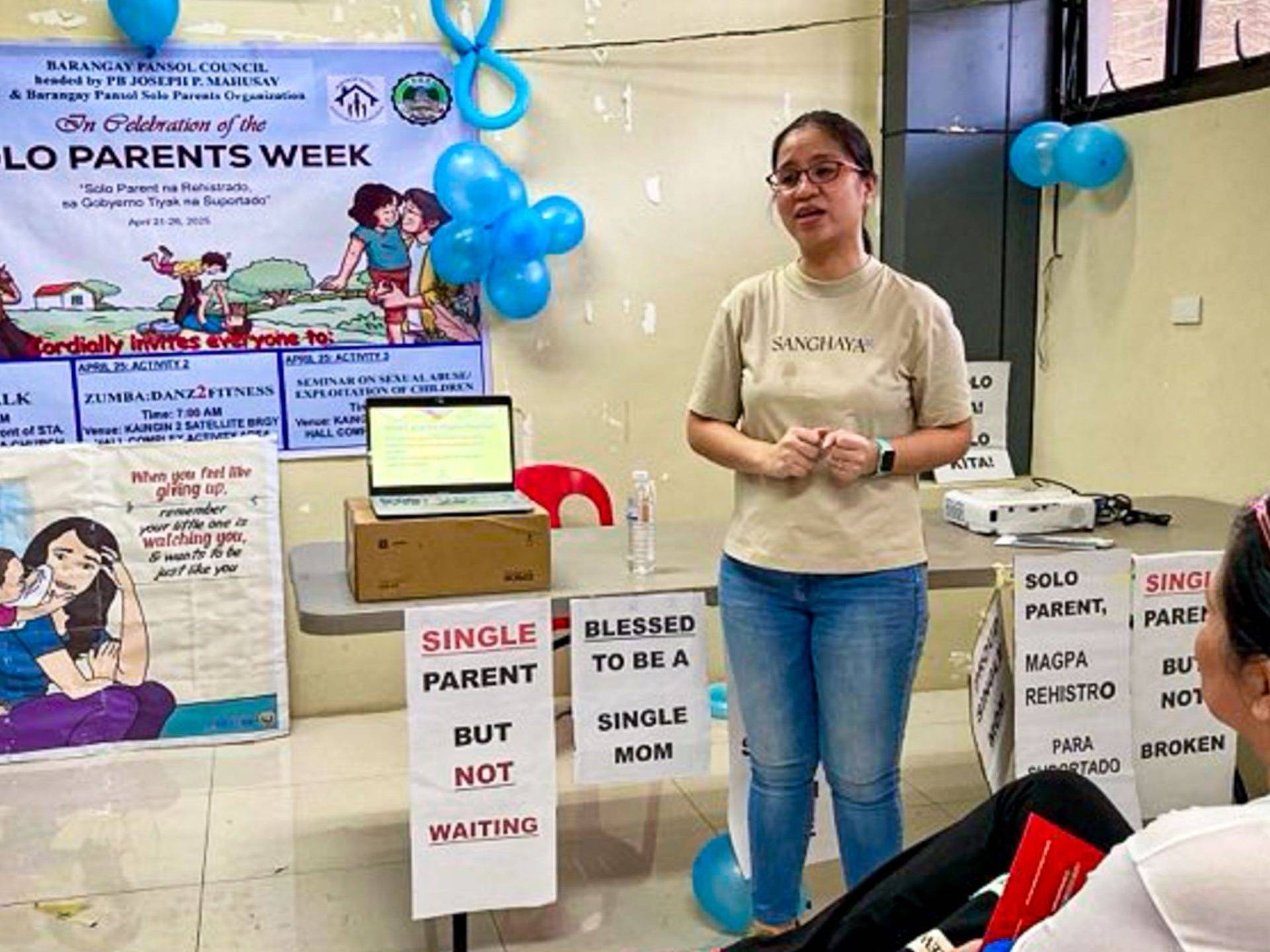 A woman poses in front of a table speaking in front of an audience in a room.