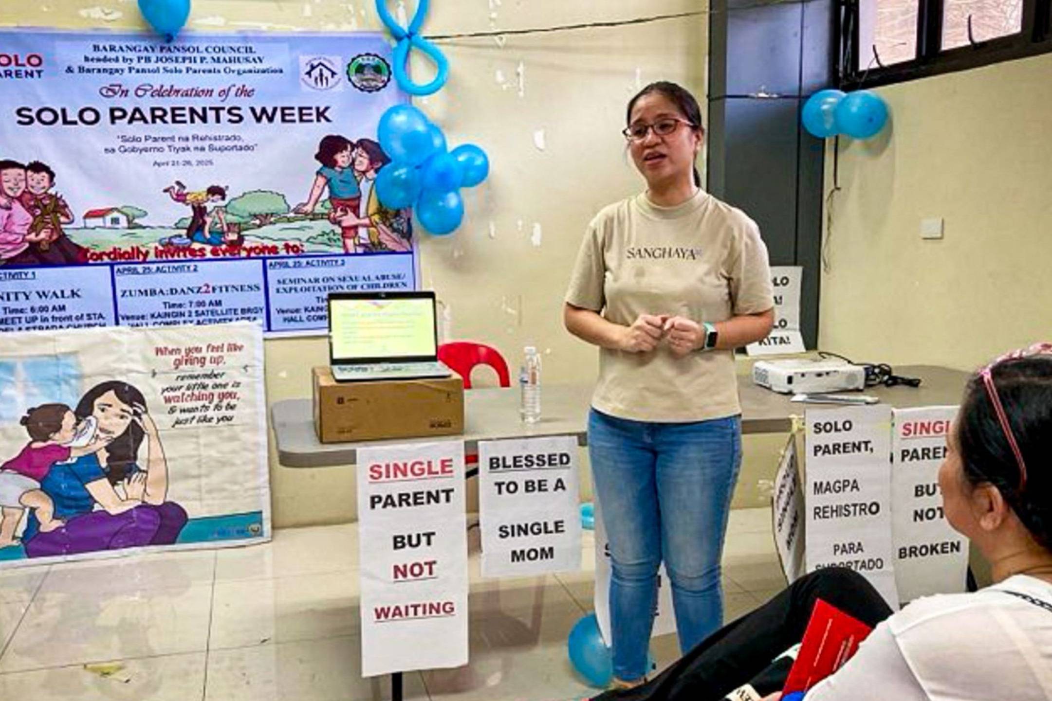 A woman poses in front of a table speaking in front of an audience in a room.